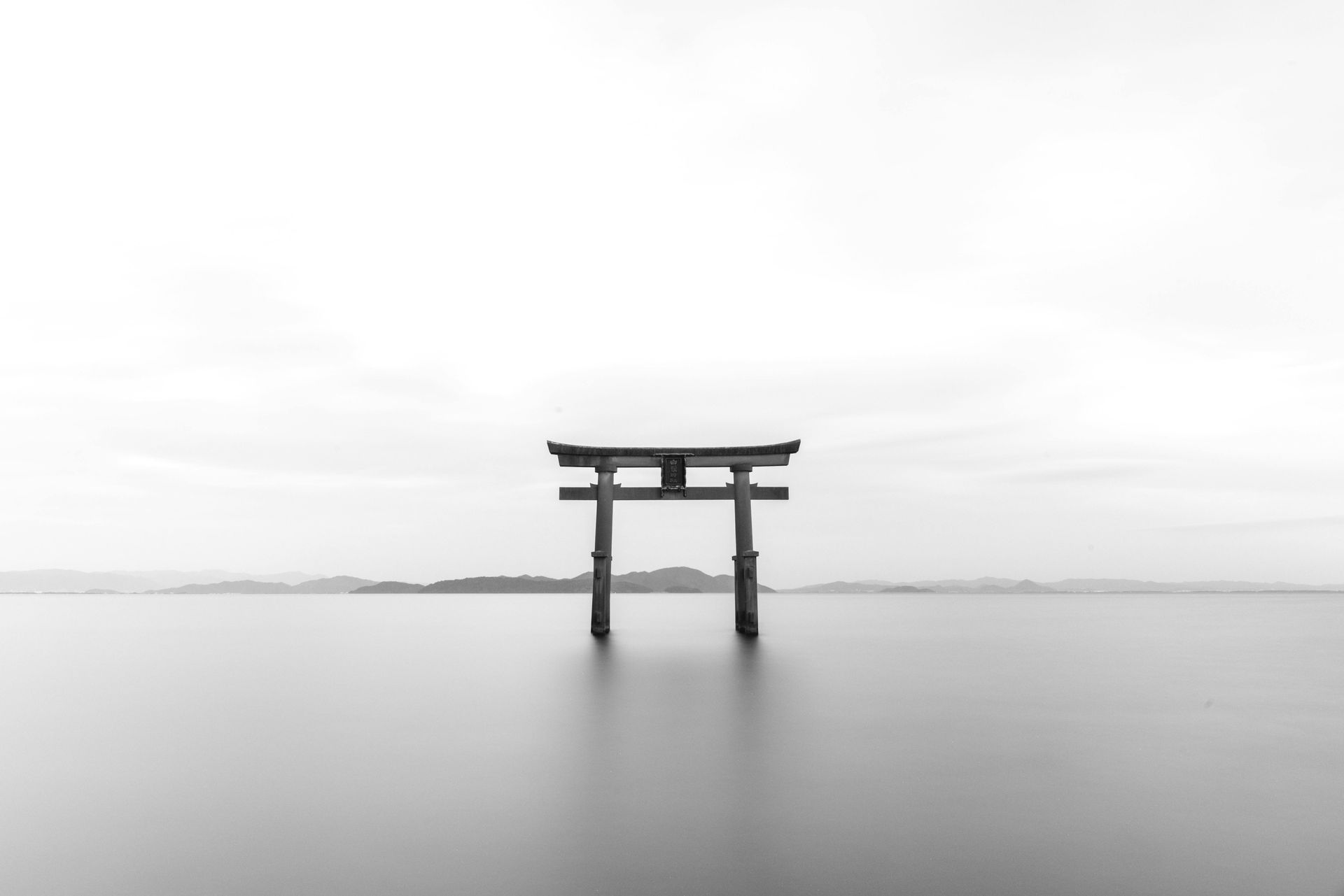 Black and white photograph of a wooden torii gate standing in tranquil water, with a hazy horizon.