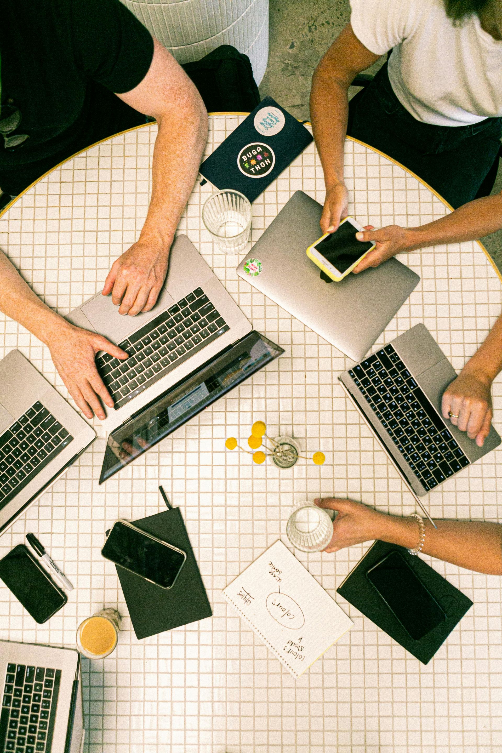 People working at a round table with laptops, phones, and papers; neutral color palette.