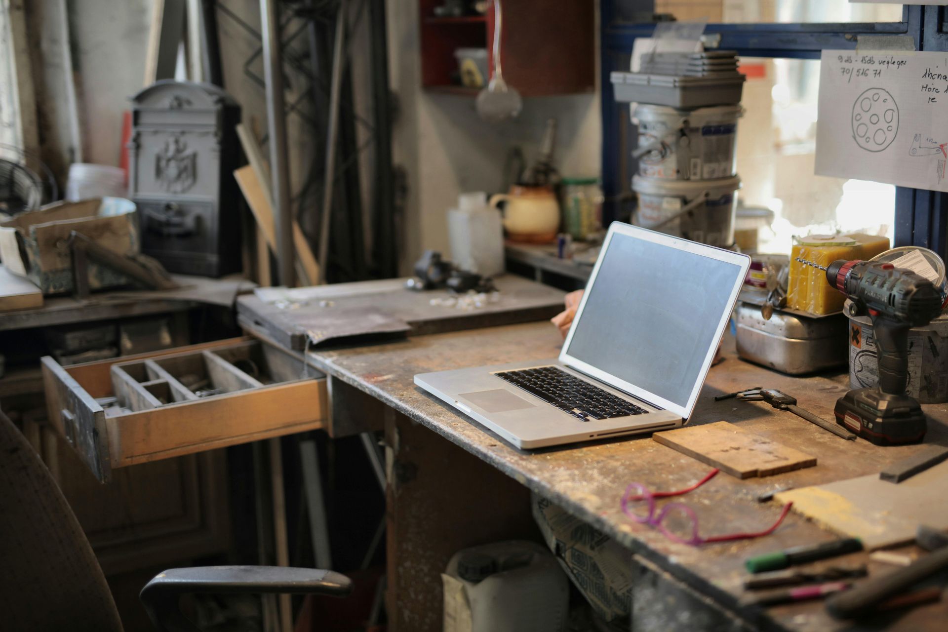 Laptop on a cluttered workbench in a workshop; tools and supplies surround it.