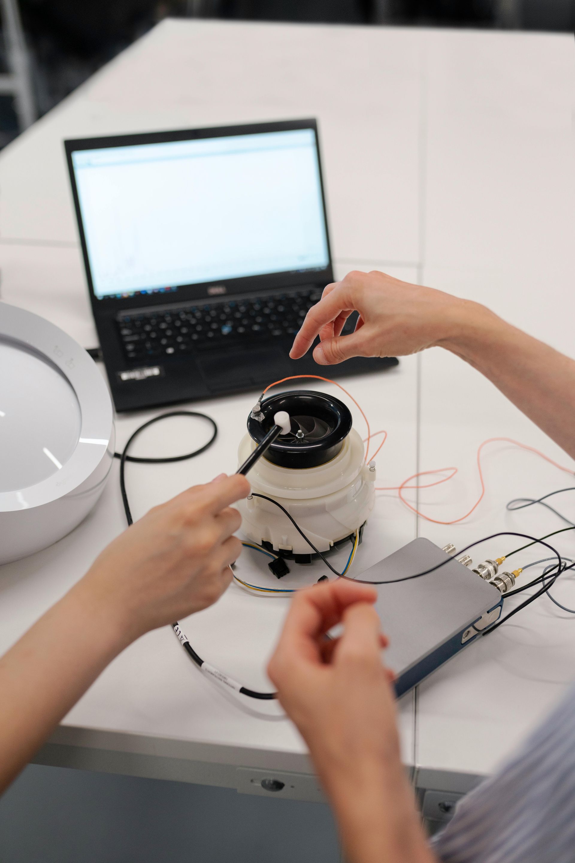 Two people working on a device with wires connected to a laptop on a white table.