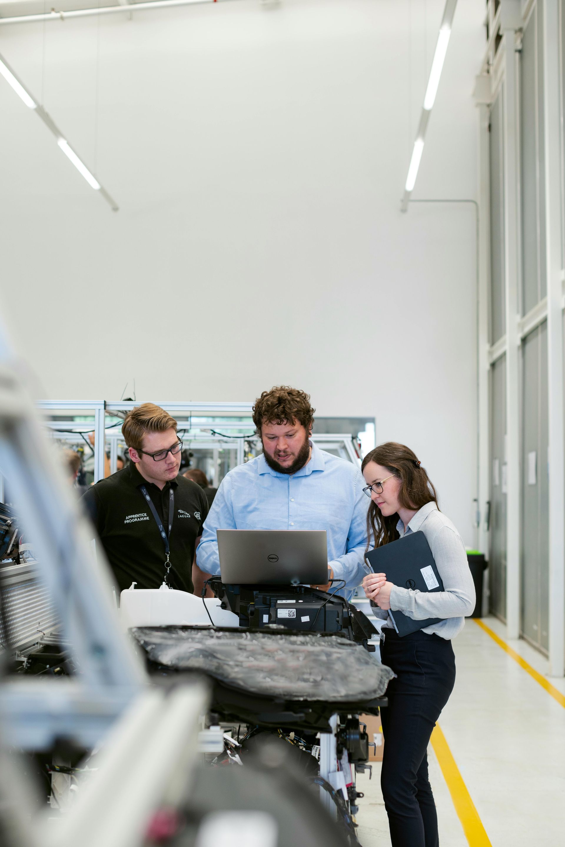 Three people looking at a laptop in a bright factory setting. A man types on the laptop, two others observe with interest.