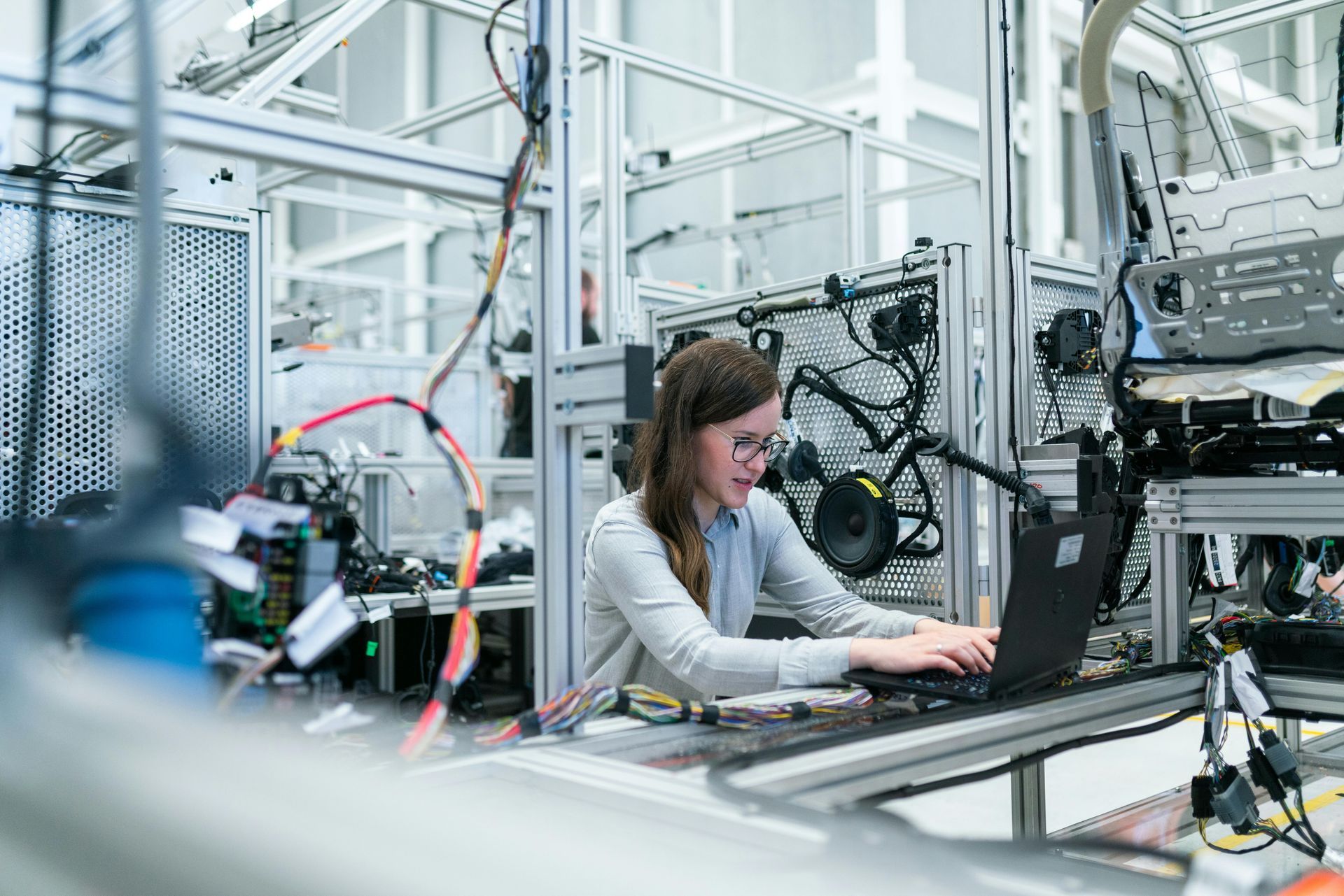 Woman in glasses using a laptop in a tech lab with machinery and wires.