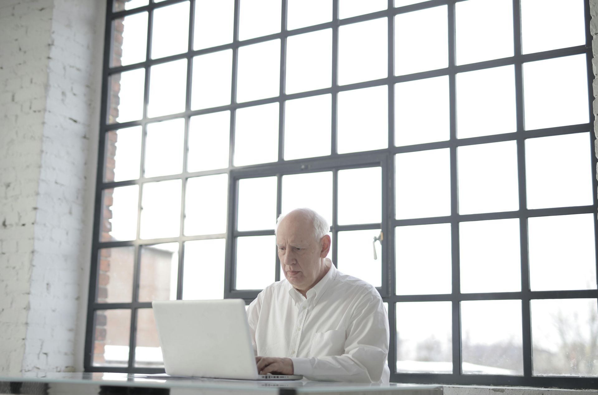 Older man in a white shirt typing on a laptop in front of a large window.