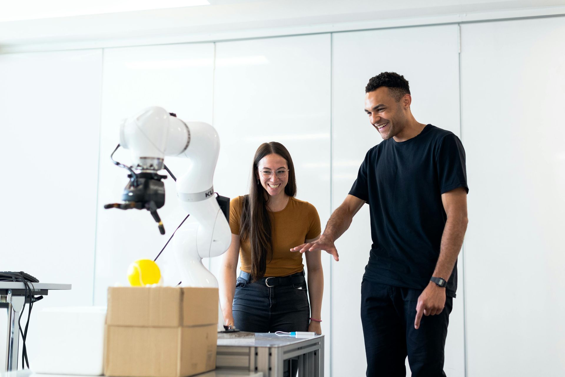 Two people watch a robotic arm place a yellow ball on a cardboard box in a lab.