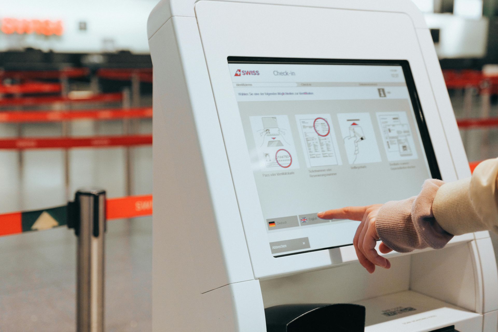 Person using airport check-in kiosk, selecting language. White machine, Swiss logo, red barriers in background.