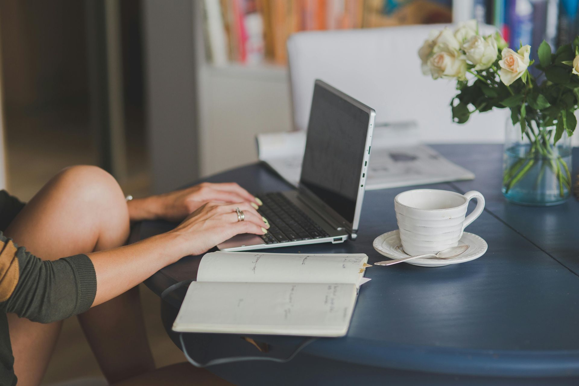Woman typing on laptop at a table with a cup of coffee, notebook, and flowers.