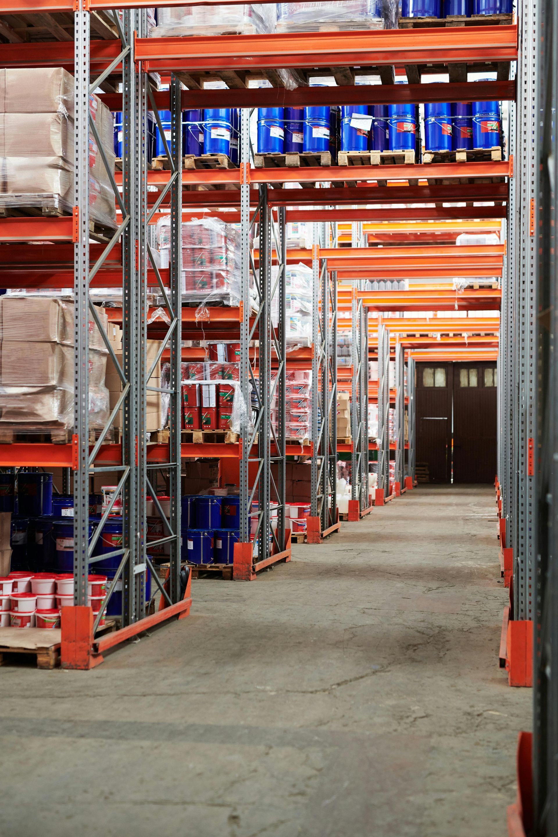 Warehouse interior with tall shelves stacked with goods, looking down the aisle to a closed door.