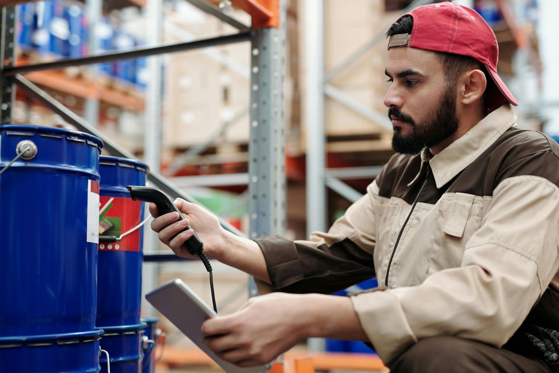 Man in work clothes and red cap scans barcode on blue container in a warehouse.