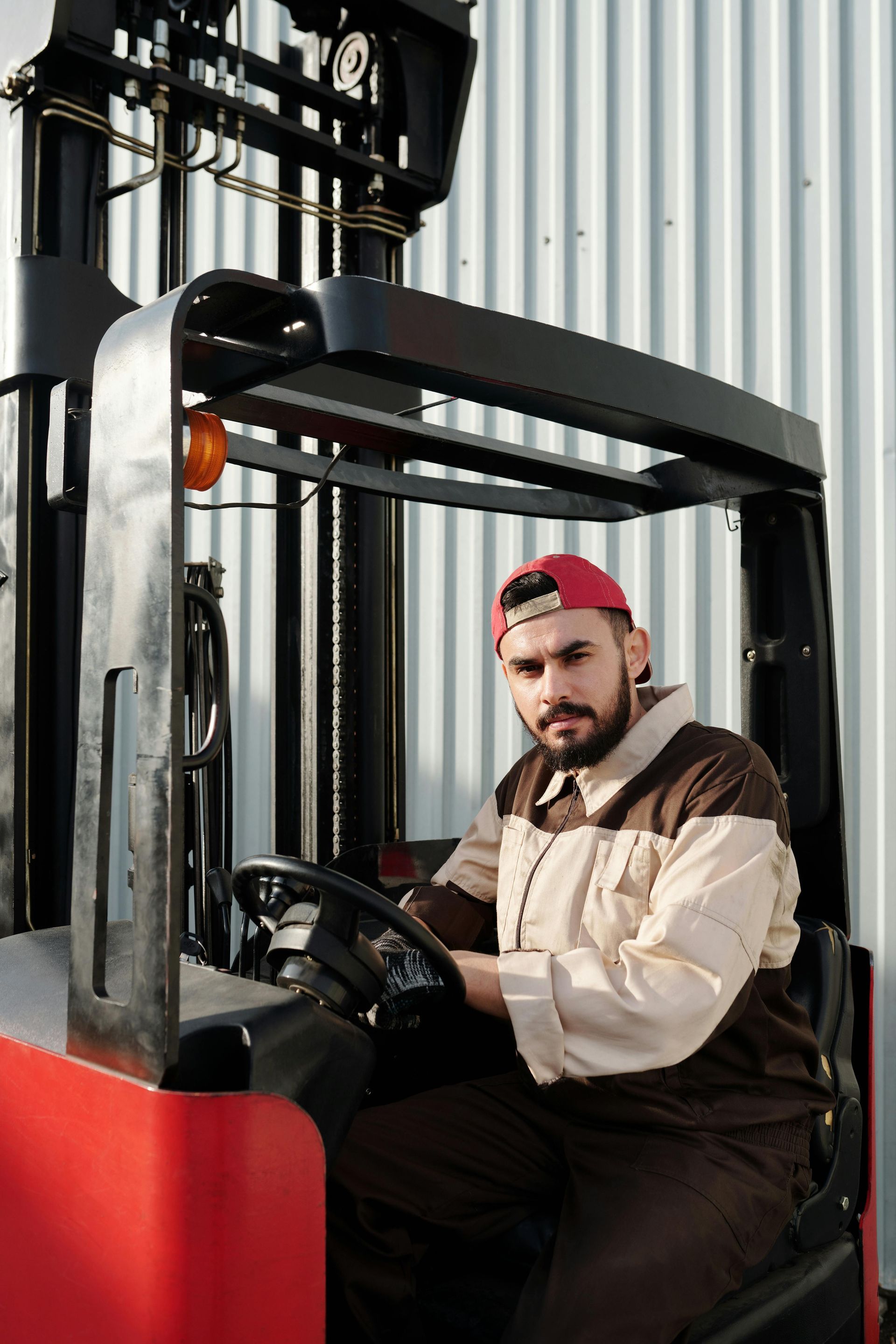 Man in work uniform operating a red forklift. Wearing a red cap, sitting in front of a corrugated wall.