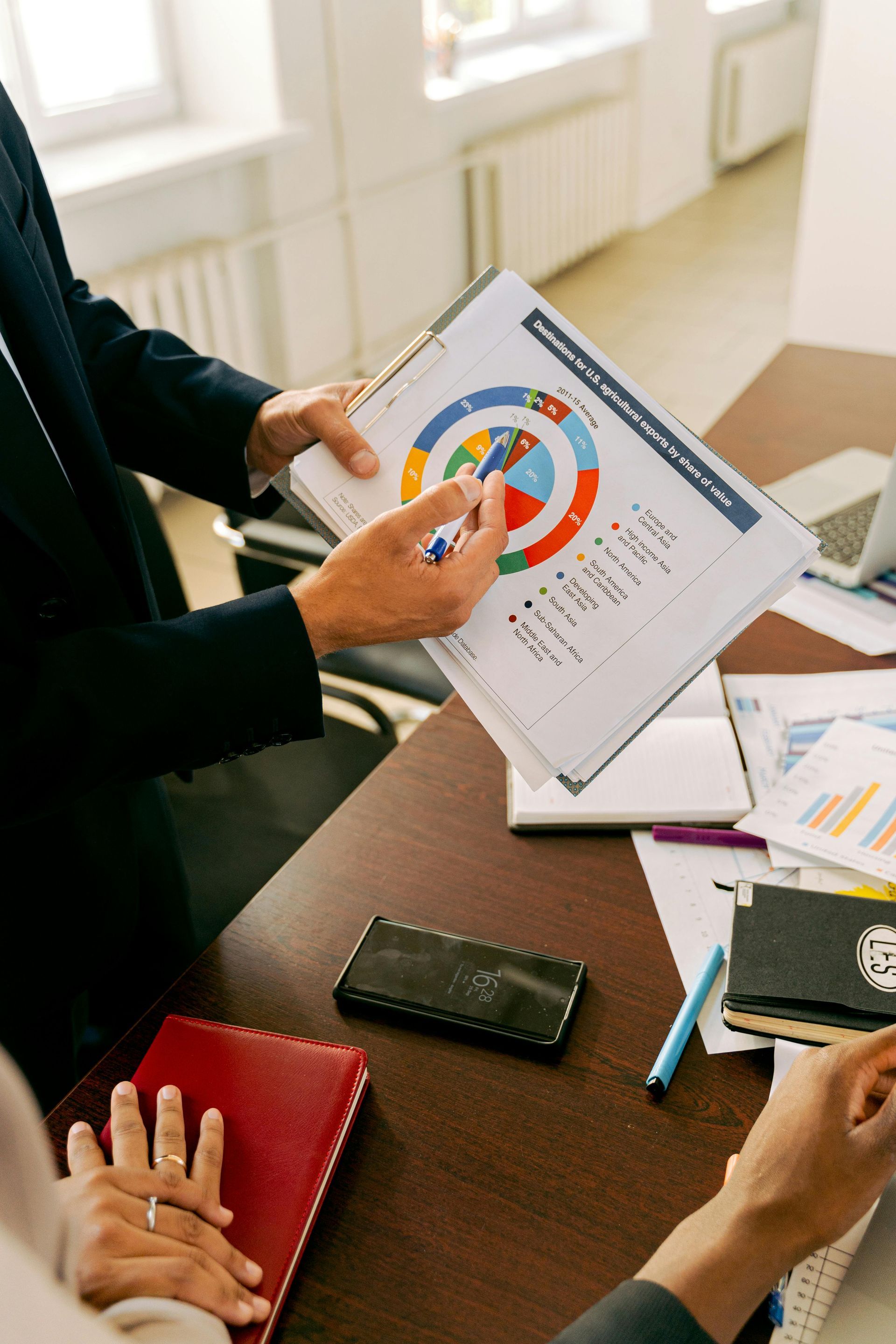 Person in suit points to a pie chart during a meeting; documents and laptop on the table.
