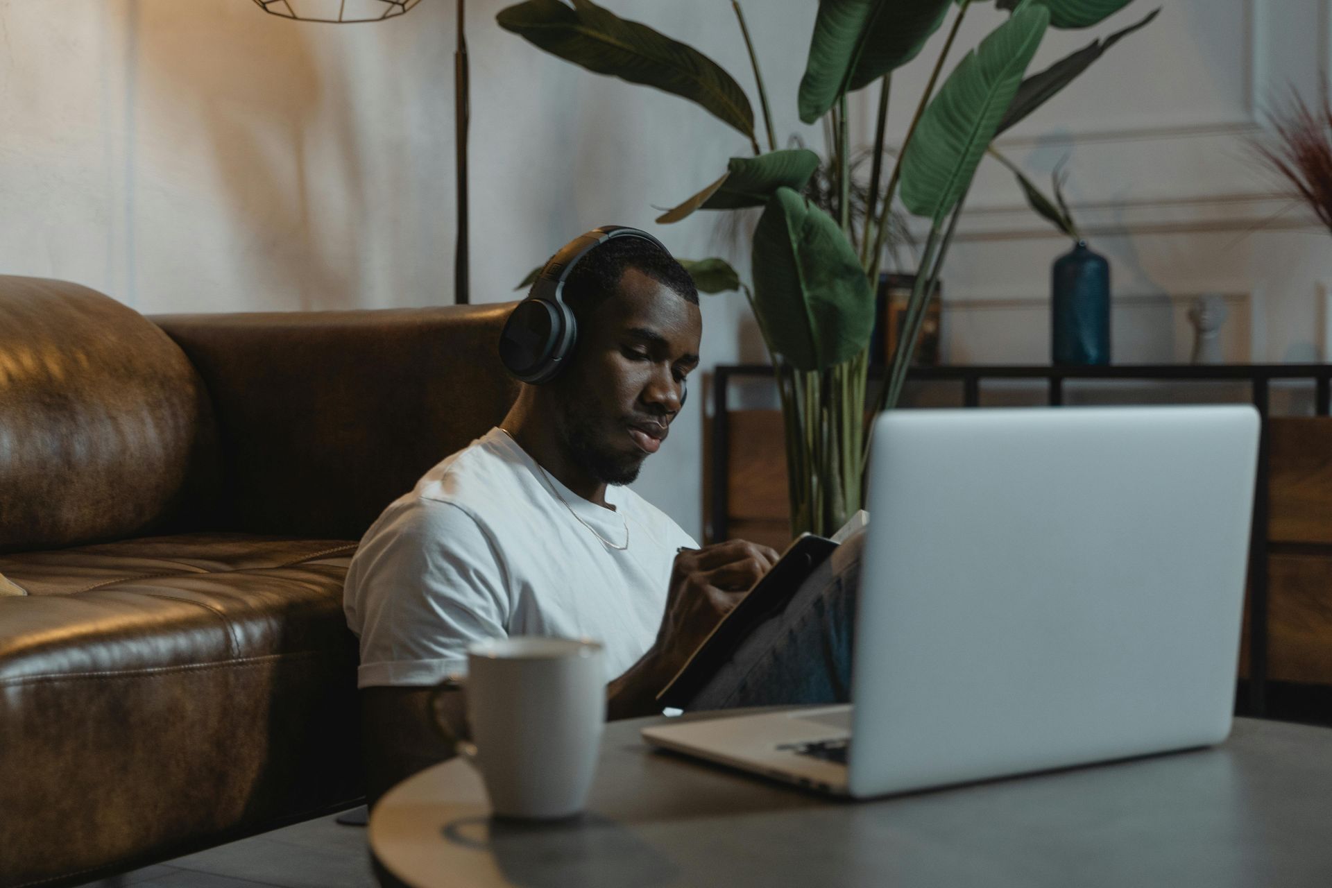 Man wearing headphones, working on a laptop while writing in a notebook, seated near a couch.