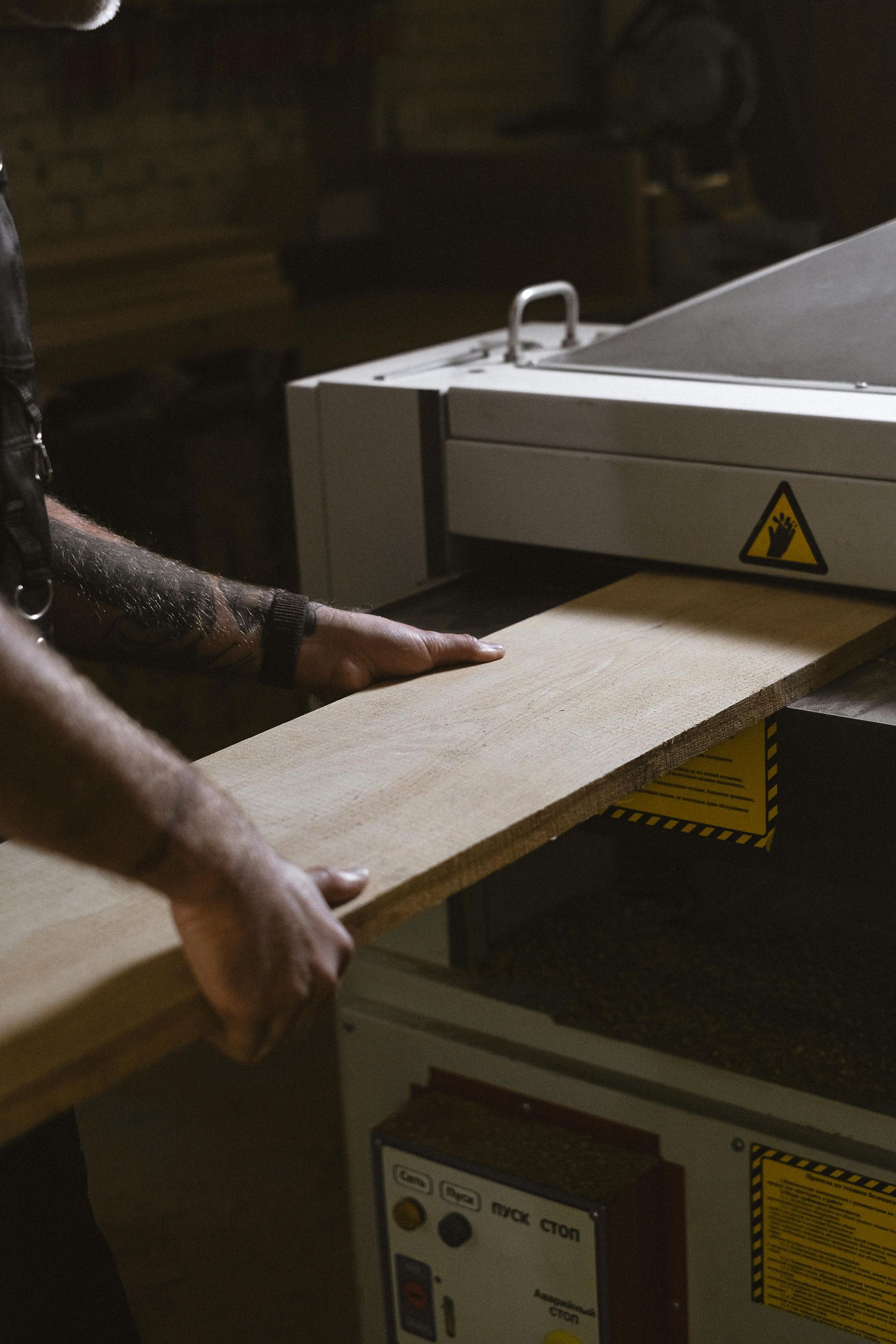 A person feeds a wooden plank into a planer machine. Industrial setting.