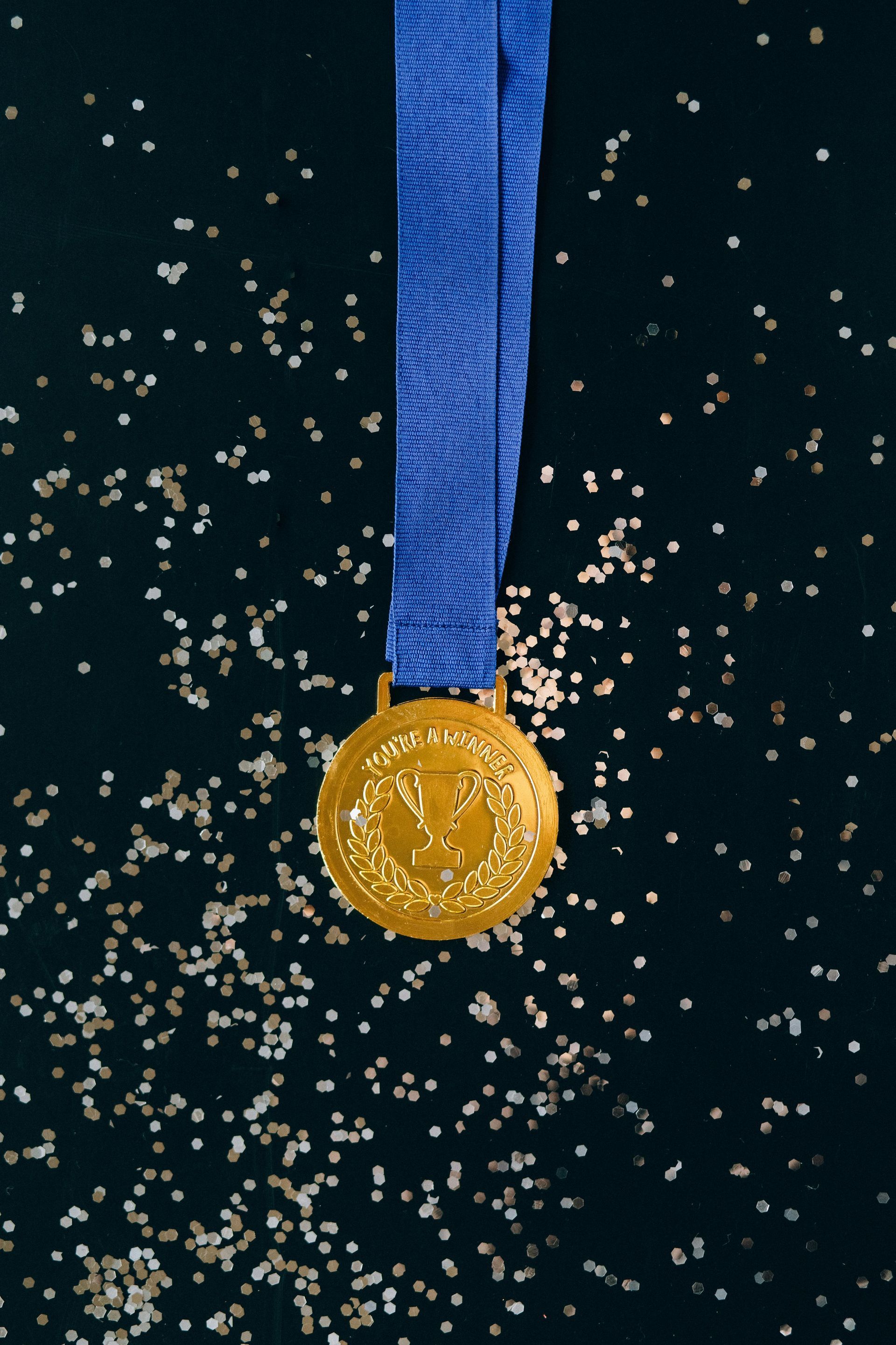Gold medal with blue ribbon, surrounded by shimmering confetti, against a dark background.