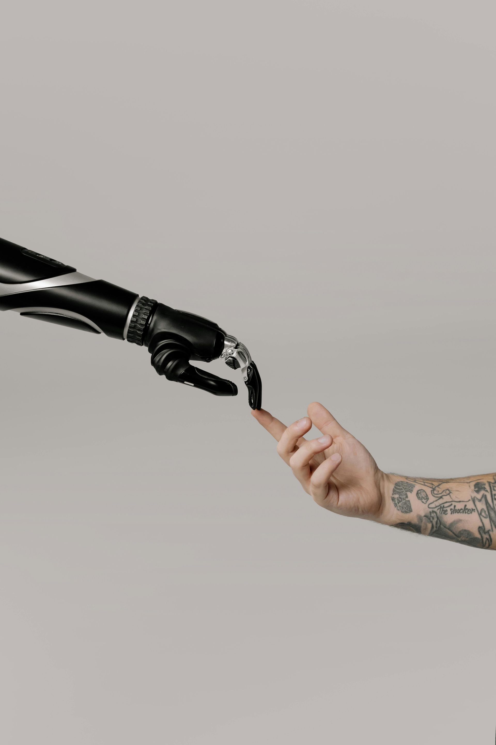 Black robotic hand touching a human finger, with tattoos. Grey background.