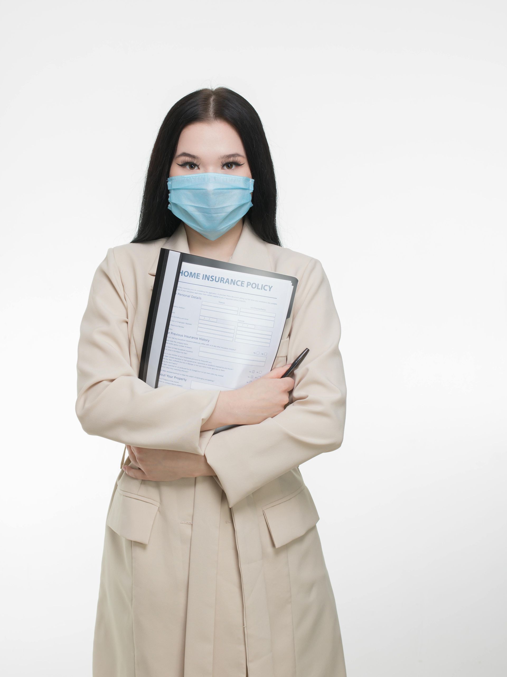 Woman in a blazer and face mask, holding a clipboard and pen, standing in front of a white background.