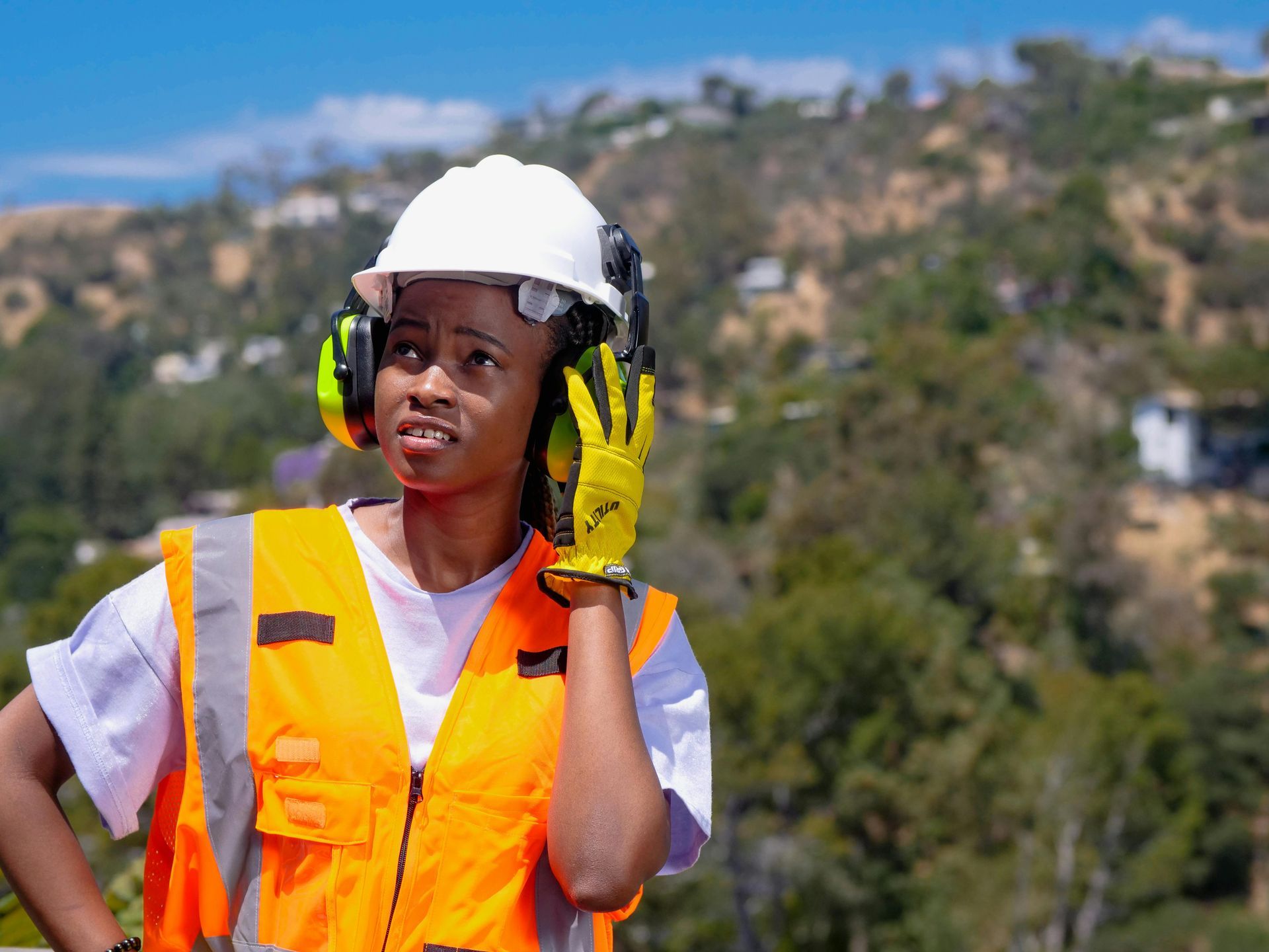 Woman in safety gear on a construction site. She's wearing a hard hat, ear protection, and safety vest, looking thoughtful.