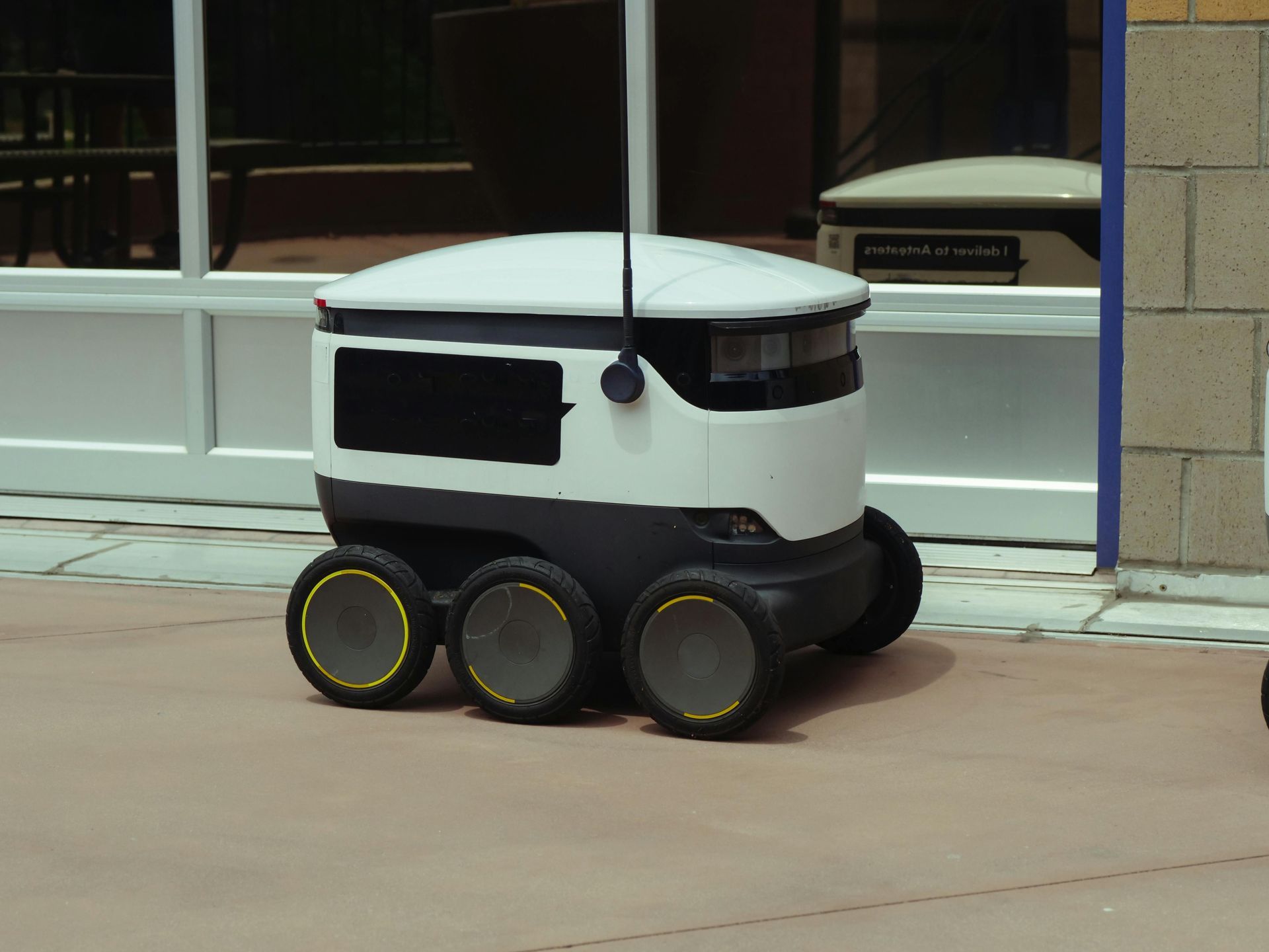 Delivery robot with a white and black body and six wheels on a sidewalk.