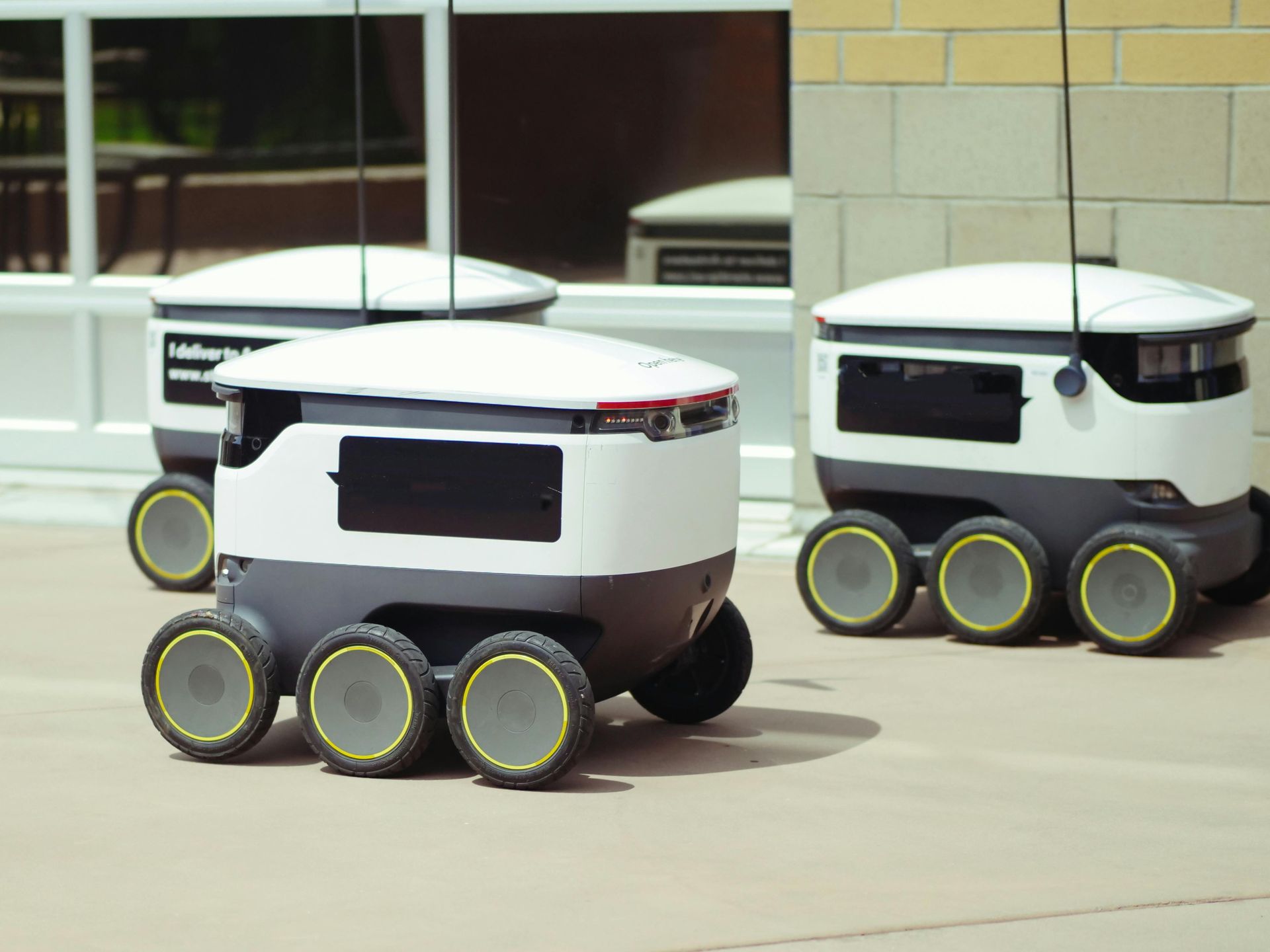 Three white and gray delivery robots with six wheels on a sidewalk near a building.