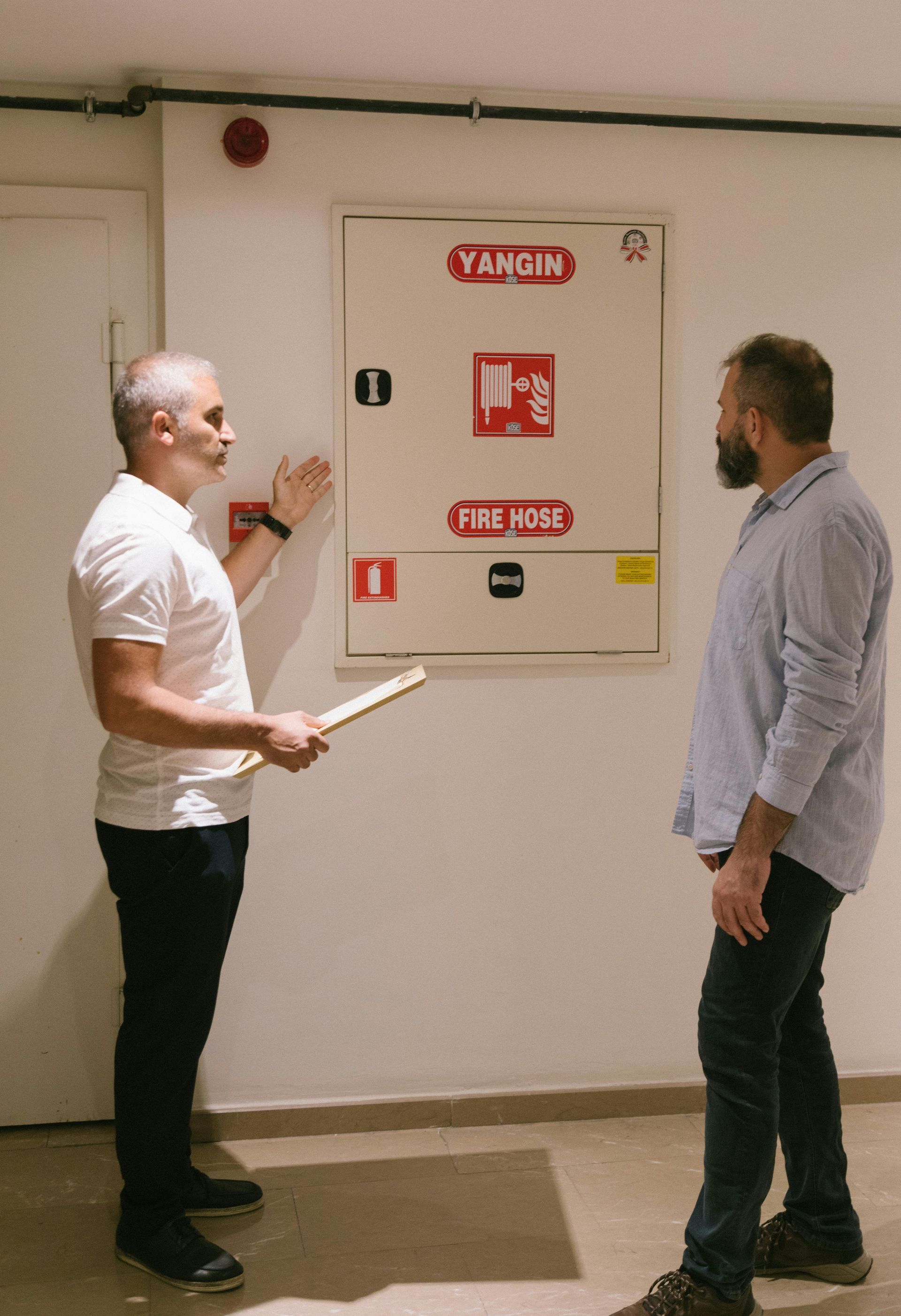Two men examining a fire safety cabinet on a white wall. One points with a stick.