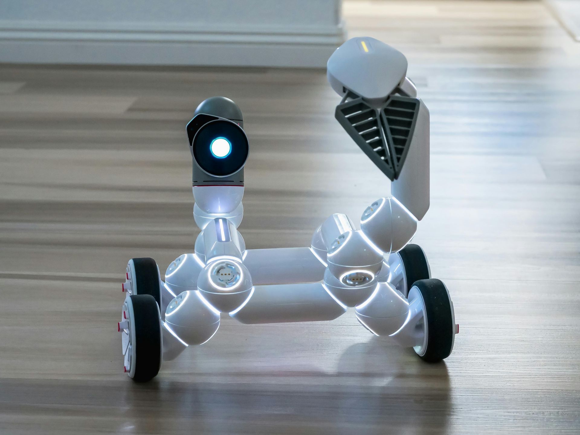White robot with glowing accents on wooden floor, tilted arm, and a camera-like head.