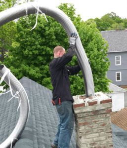 A man is standing on top of a roof holding a hose attached to a chimney.