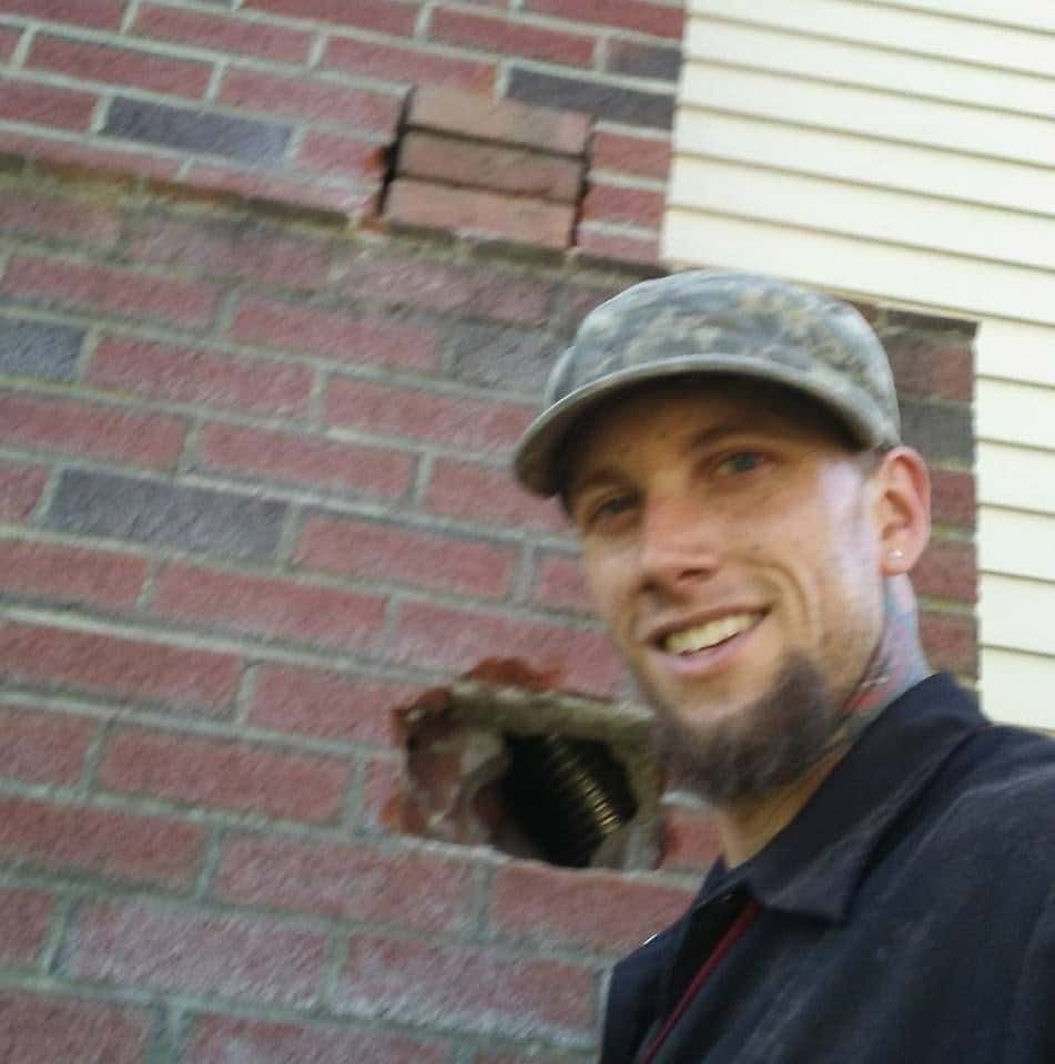 A man wearing a hat is smiling in front of a brick wall
