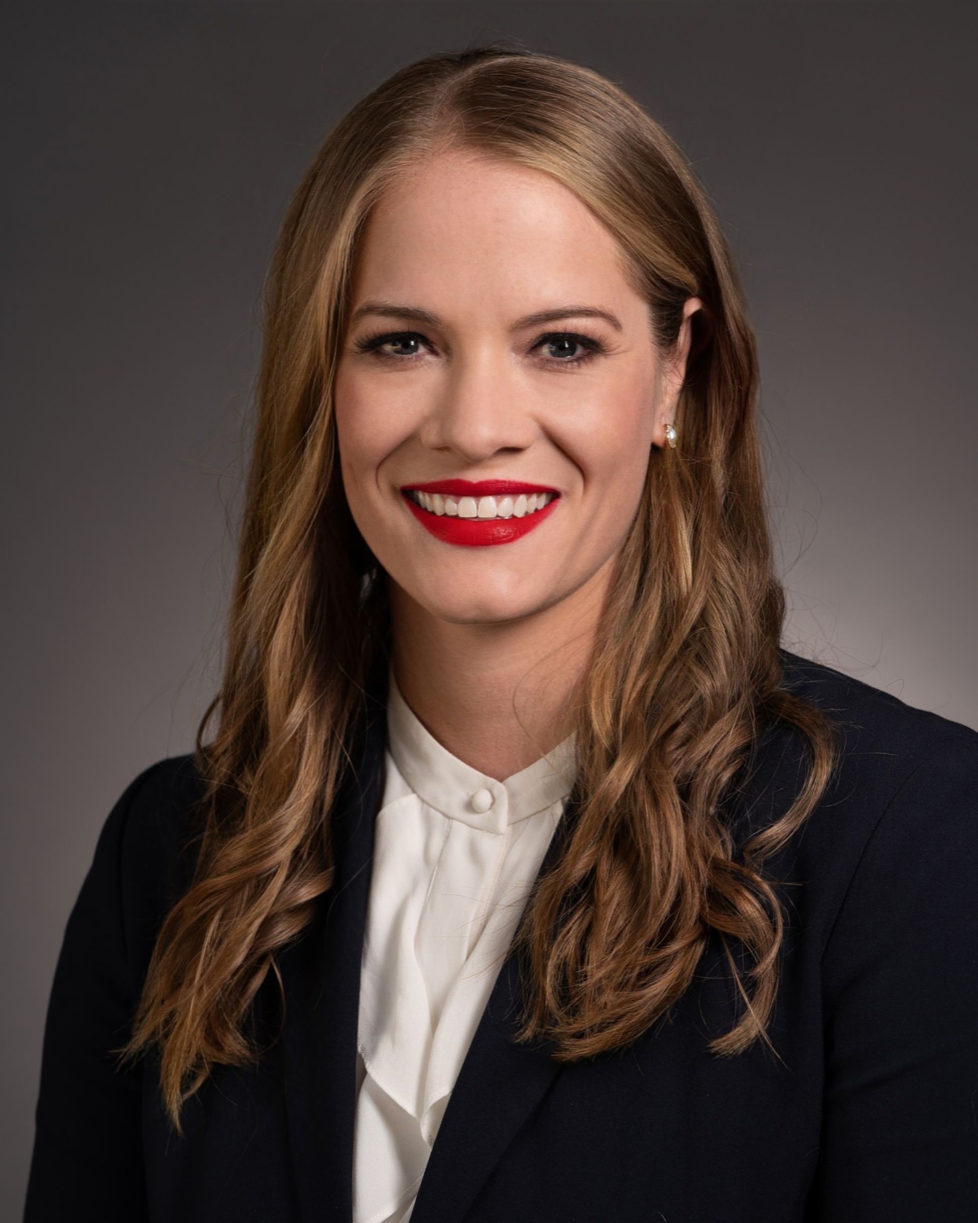 Woman with long wavy brown hair, red lipstick, and a navy blazer smiles at the camera.