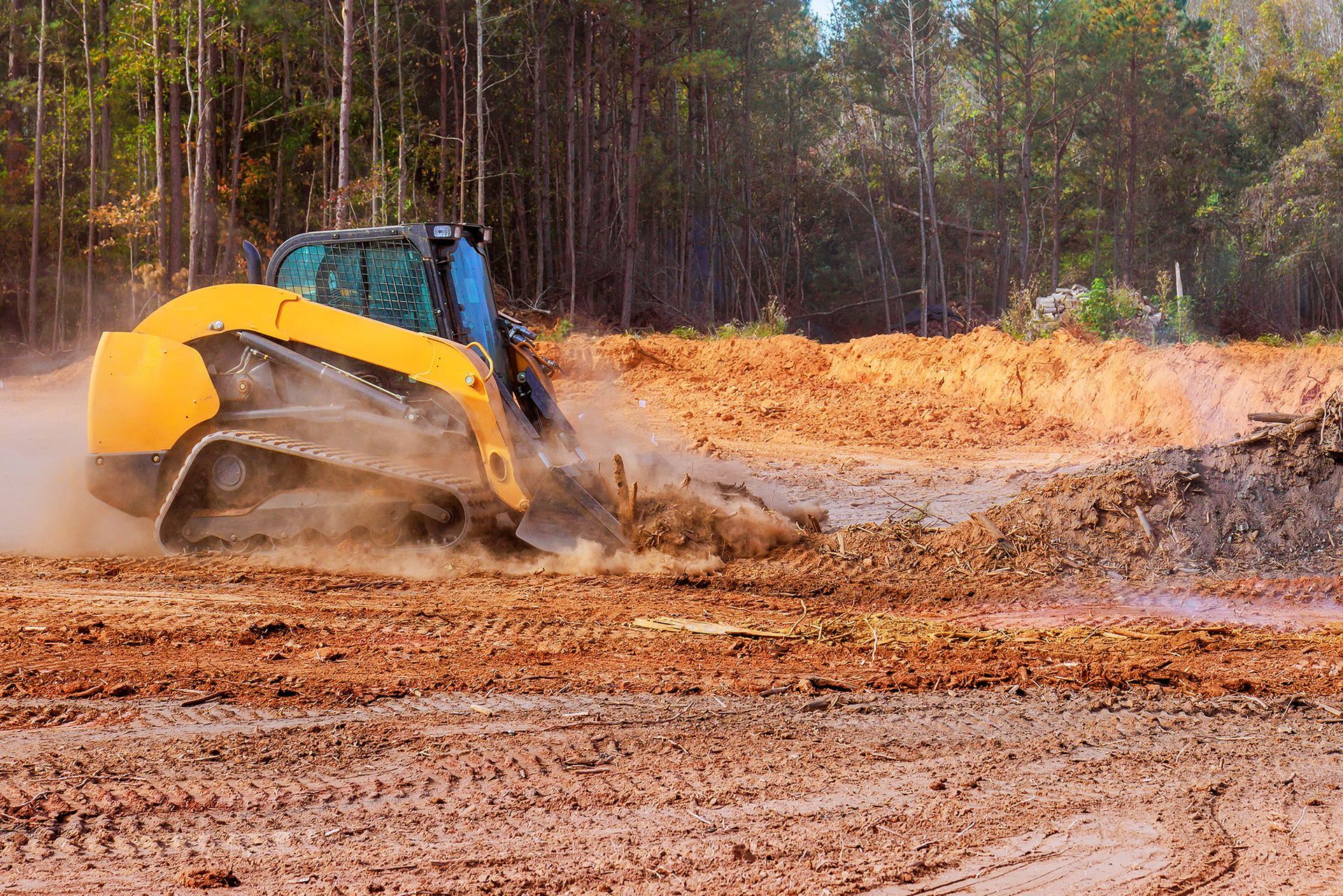 Yellow and blue skid-steer loader moving dirt in a construction site with a forest in the background.