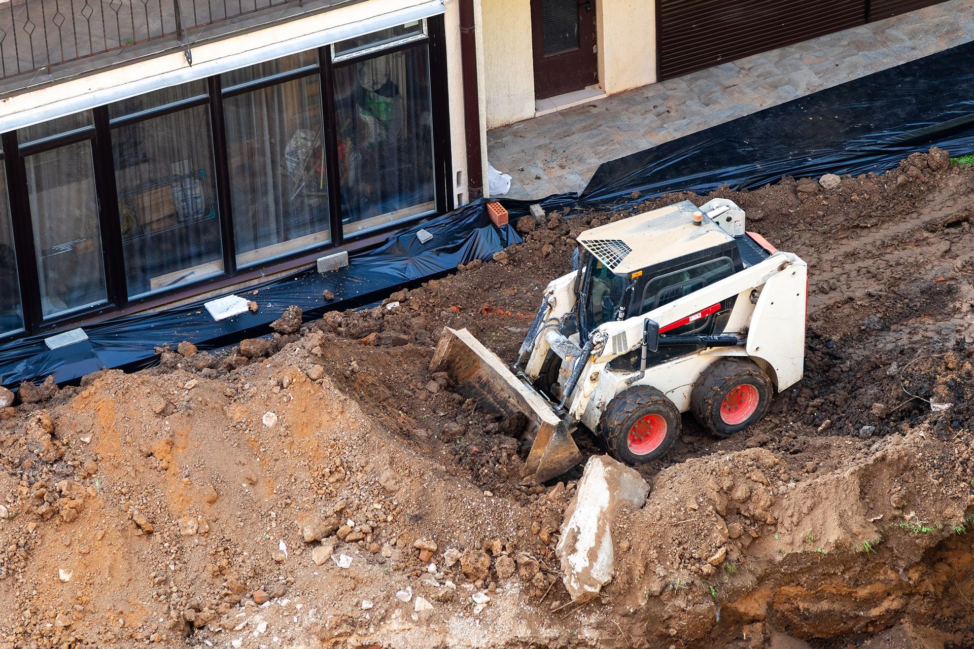 Small white Bobcat skid-steer loader pushing dirt near a building with black windows.