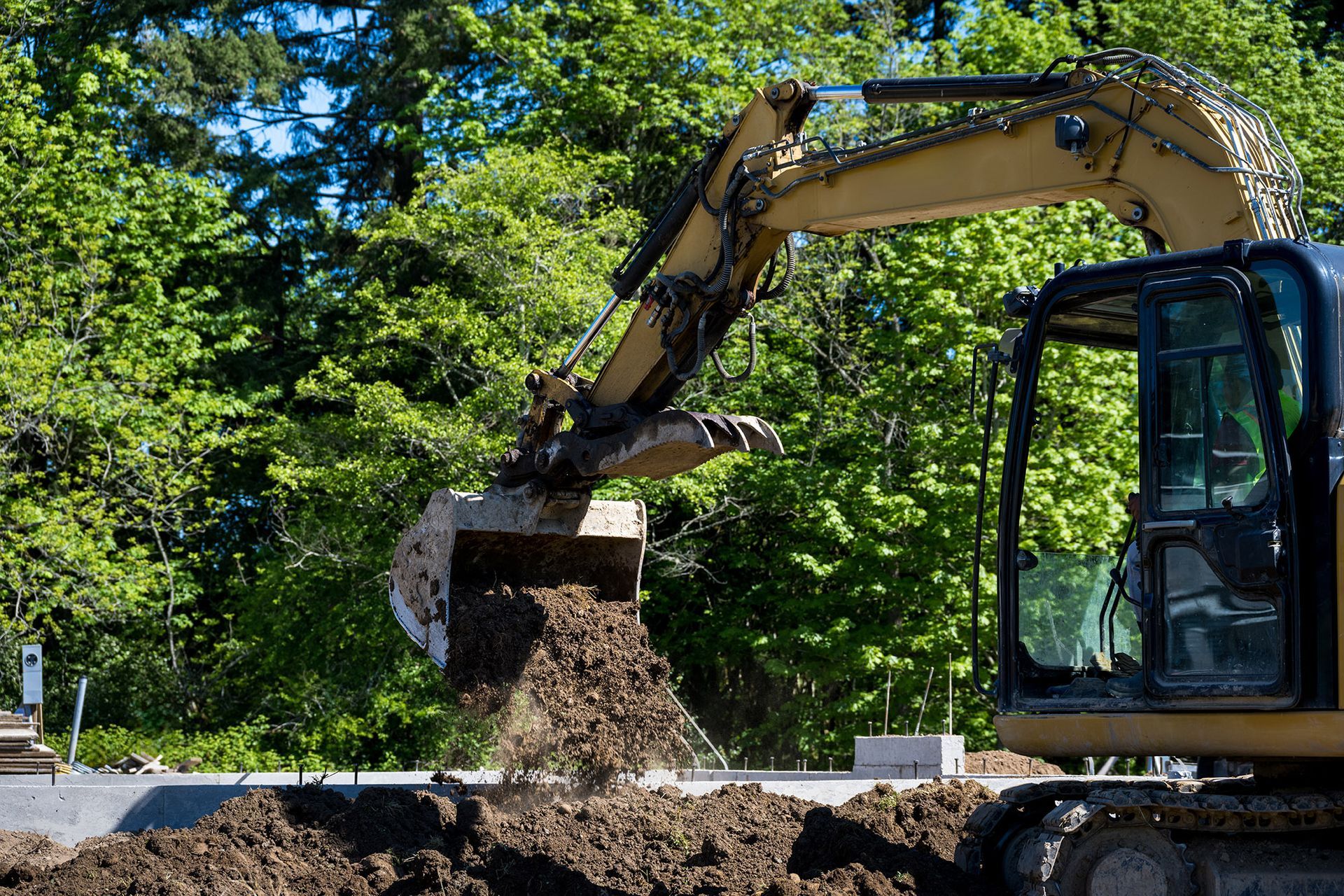 Yellow excavator dumping soil near a structure, green trees in background.