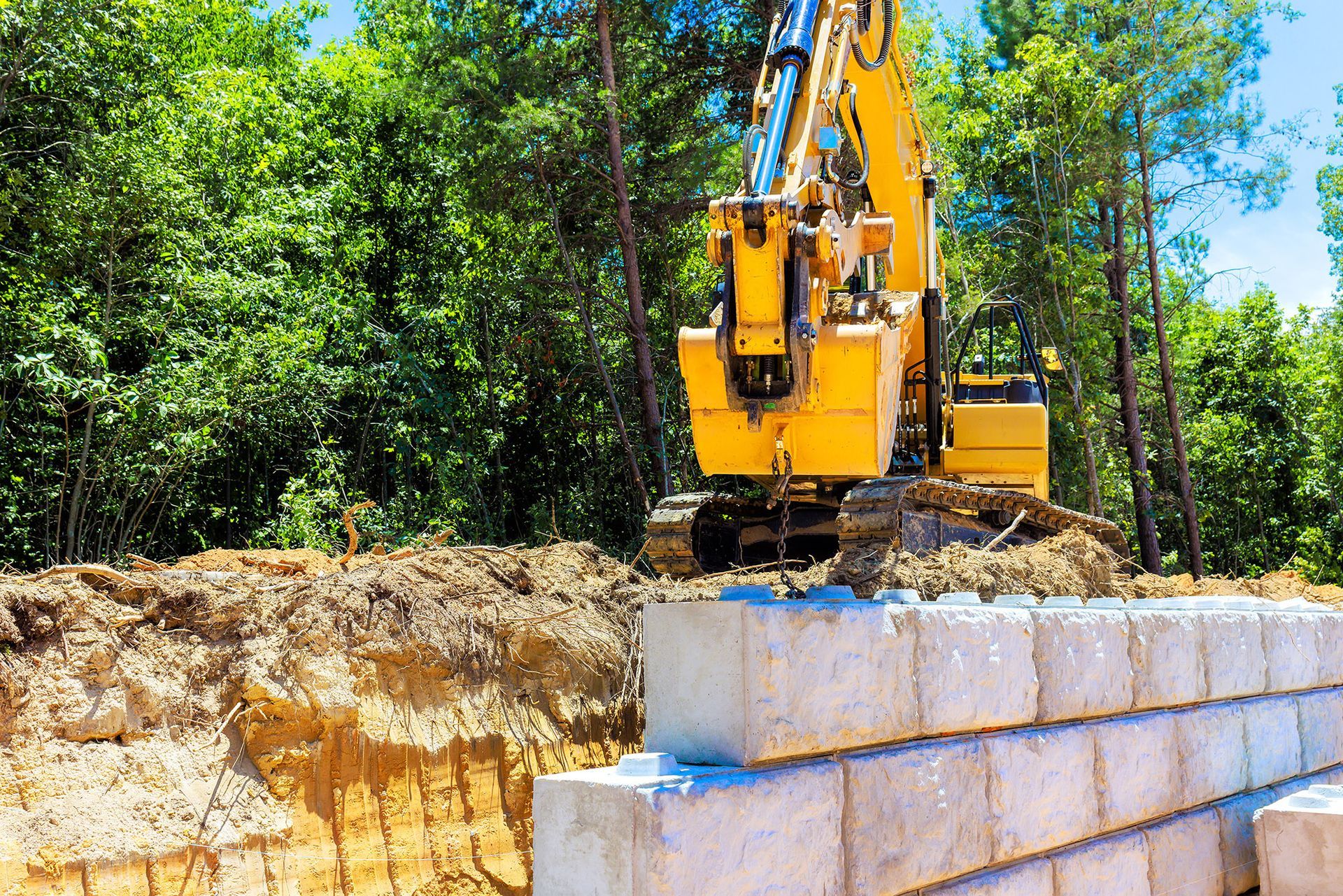 Yellow excavator building concrete block retaining wall near trees.