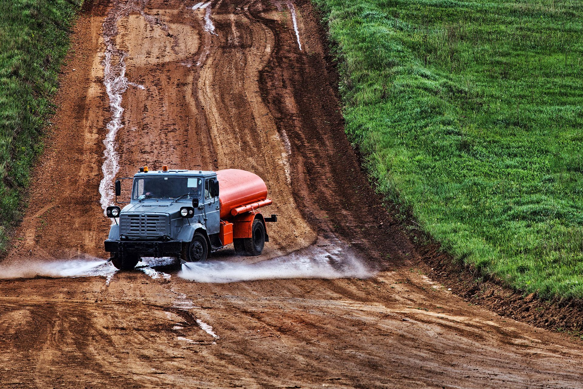 Tanker truck drives through muddy terrain, splashing water.