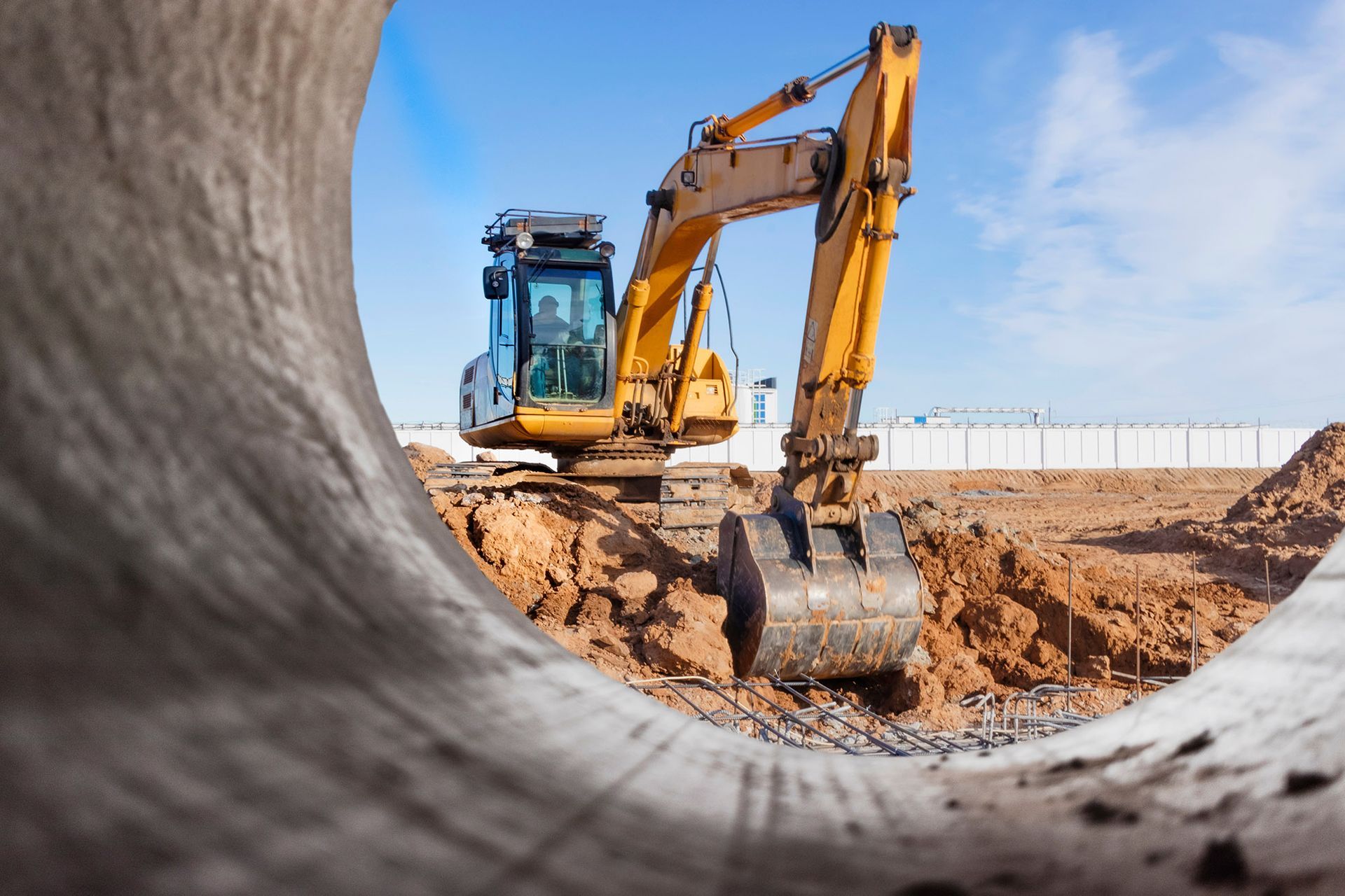 Yellow excavator digging earth through a concrete pipe, under a blue sky.