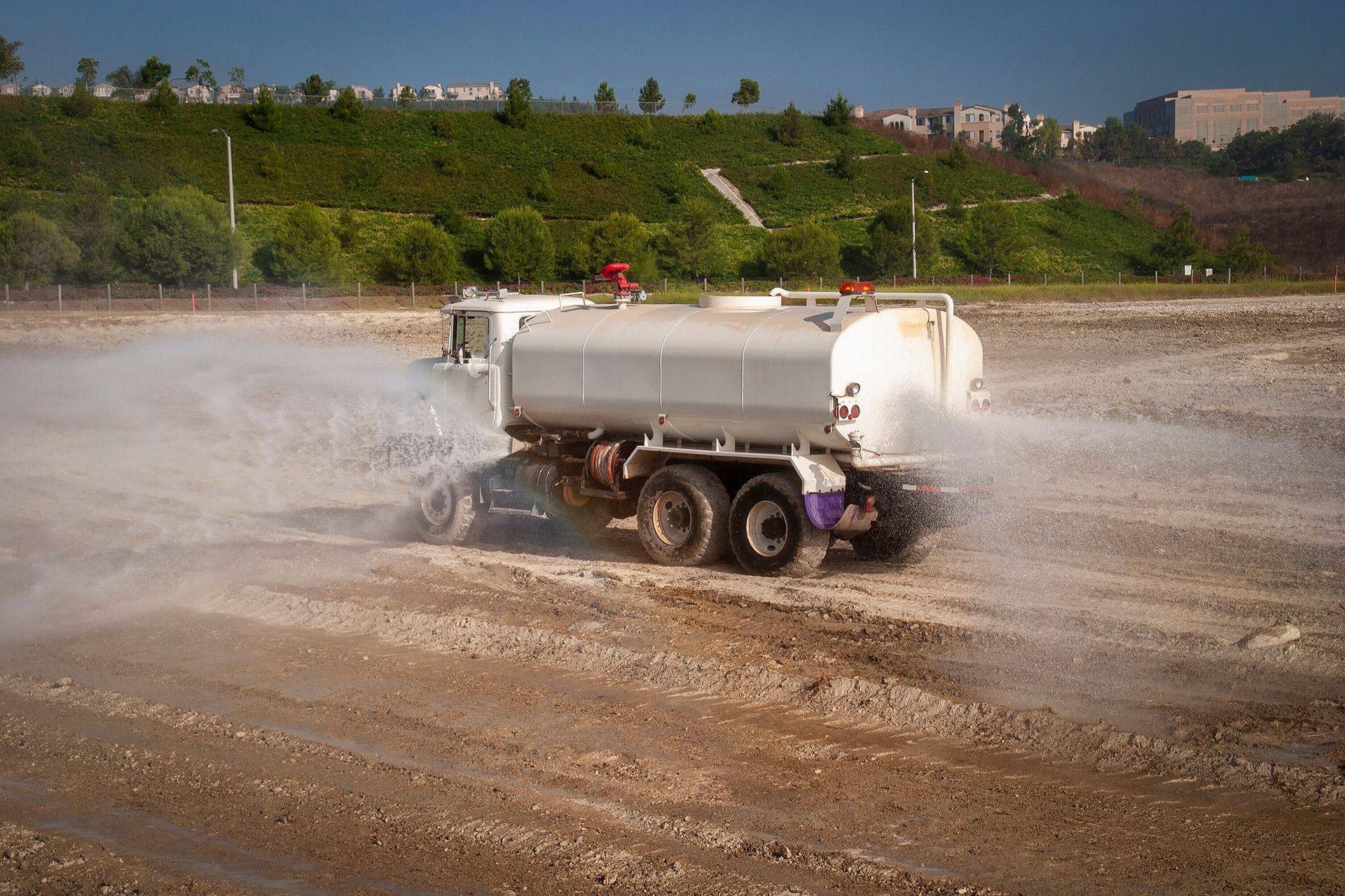 Water truck spraying water on a dry, dirt field; green hillside in background.