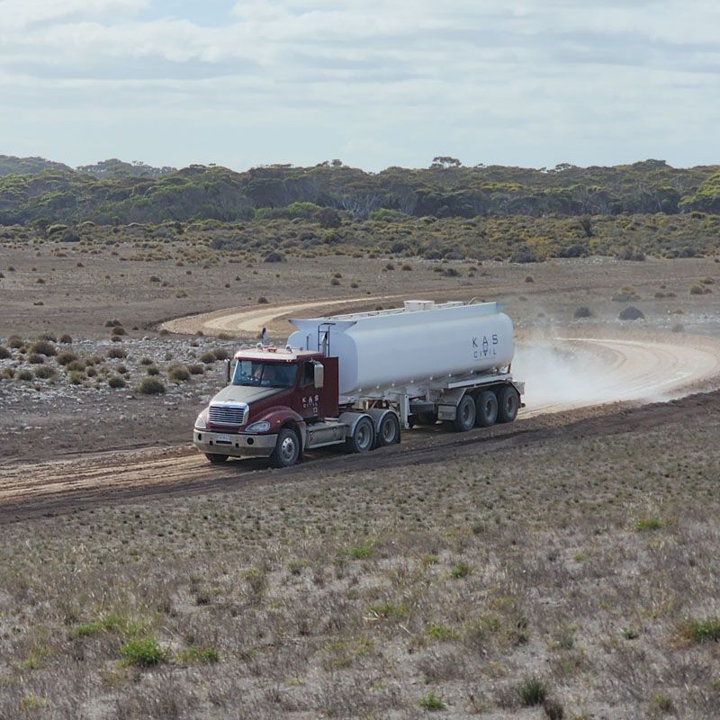 Semi-truck hauling a tanker travels on a dirt road, kicking up dust in a dry, rural landscape.