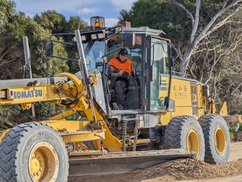 Yellow Komatsu motor grader with operator on a dirt road, trees in the background, sunny day.