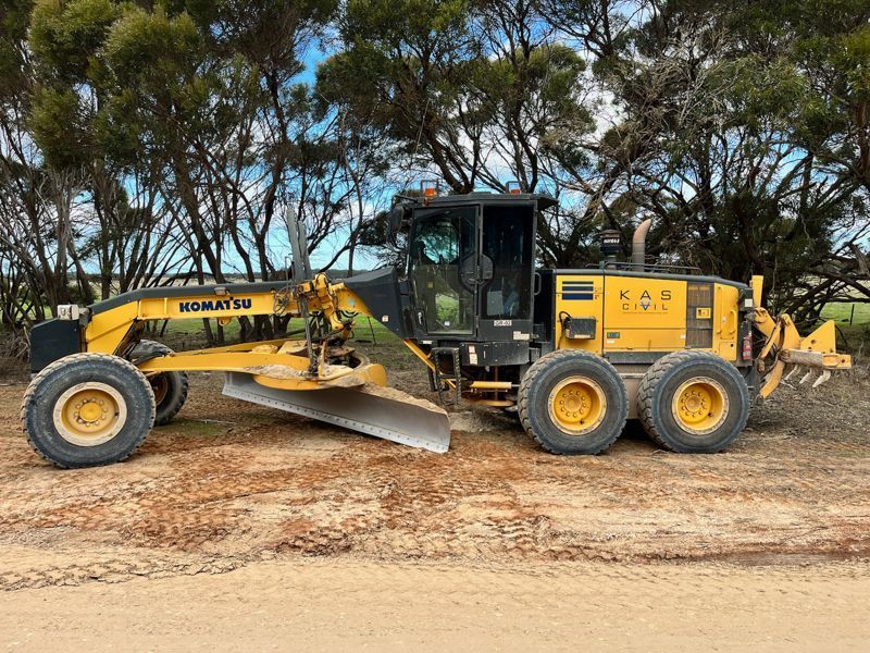 Yellow Komatsu grader on a dirt road, next to trees.