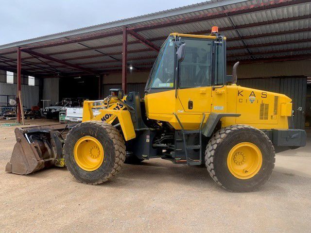 Yellow KAS wheel loader parked under a metal roof structure with a loading bucket.