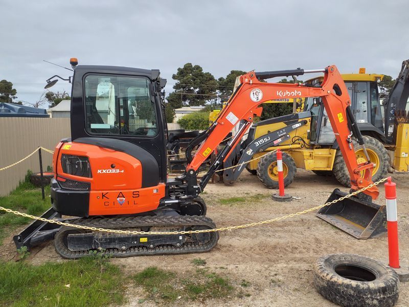 Orange Kubota excavator on dirt with a yellow loader in the background.