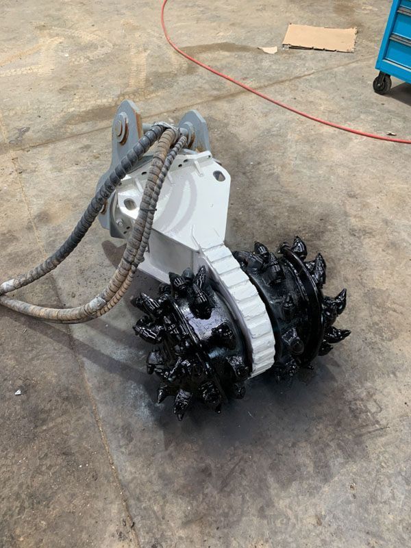 White and black mining attachment with rotating spiked wheels, connected to hoses, lying on concrete.