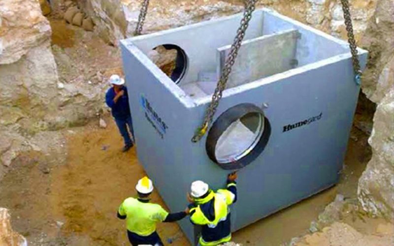 A concrete drainage structure being lowered into a trench by a crane, with construction workers nearby.