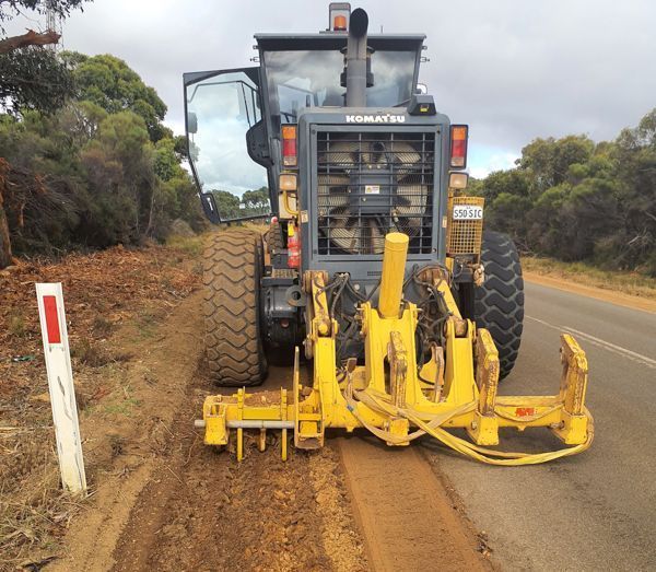 Yellow Komatsu grader on roadside, smoothing the shoulder. Gray and brown colors dominate the scene.