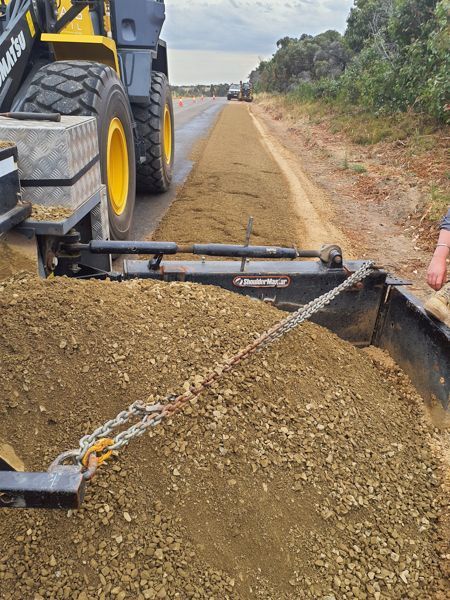 Road construction: yellow loader pulls a spreader, depositing gravel on a dirt road.