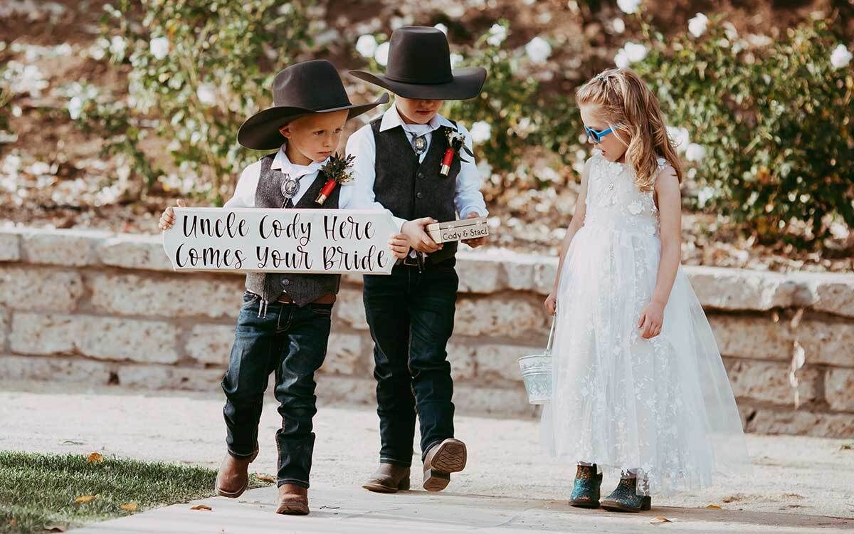 The ring bearer and flower girl are wearing cowboy hats and holding a sign.