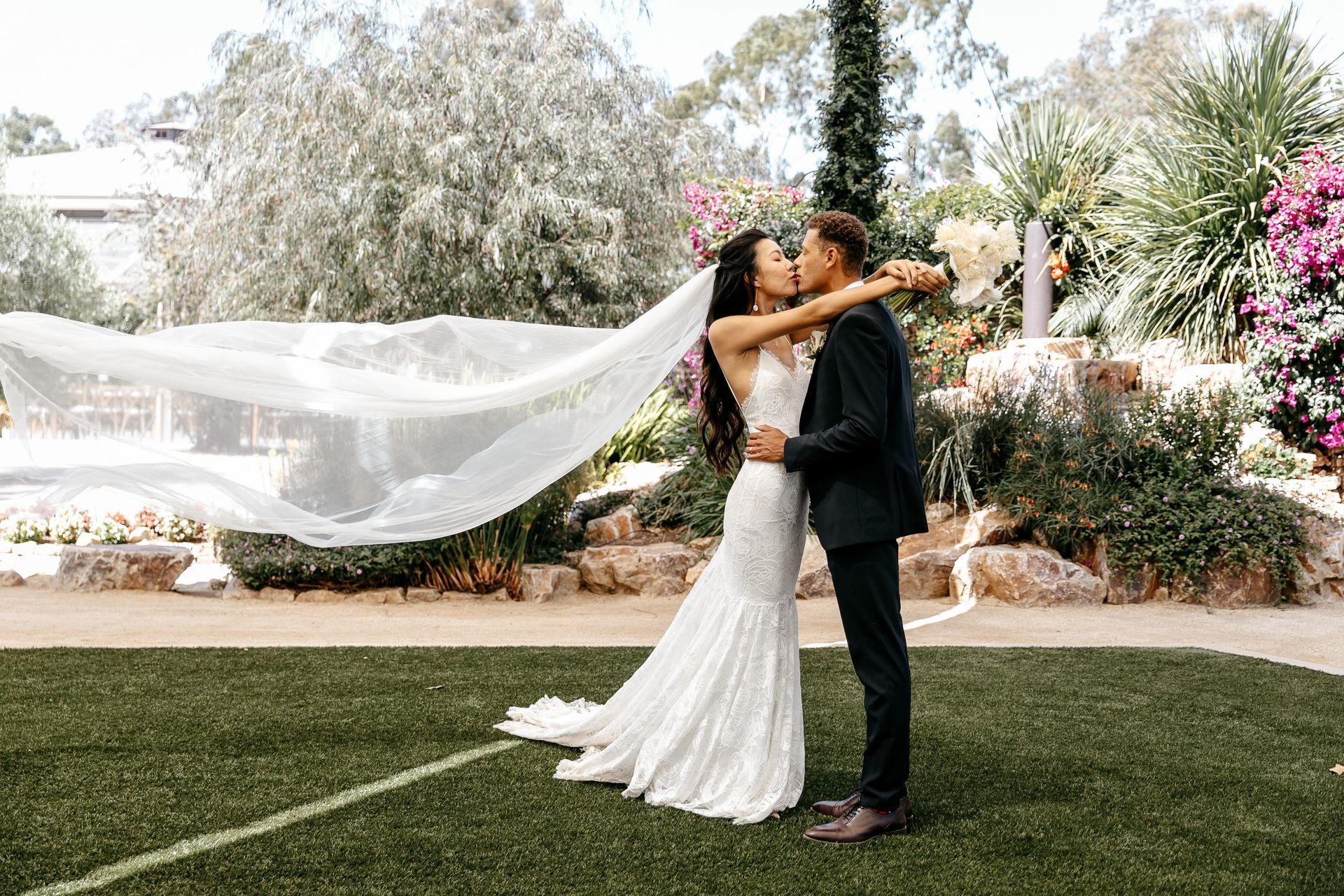 A bride and groom are kissing in a field with a veil blowing in the wind.