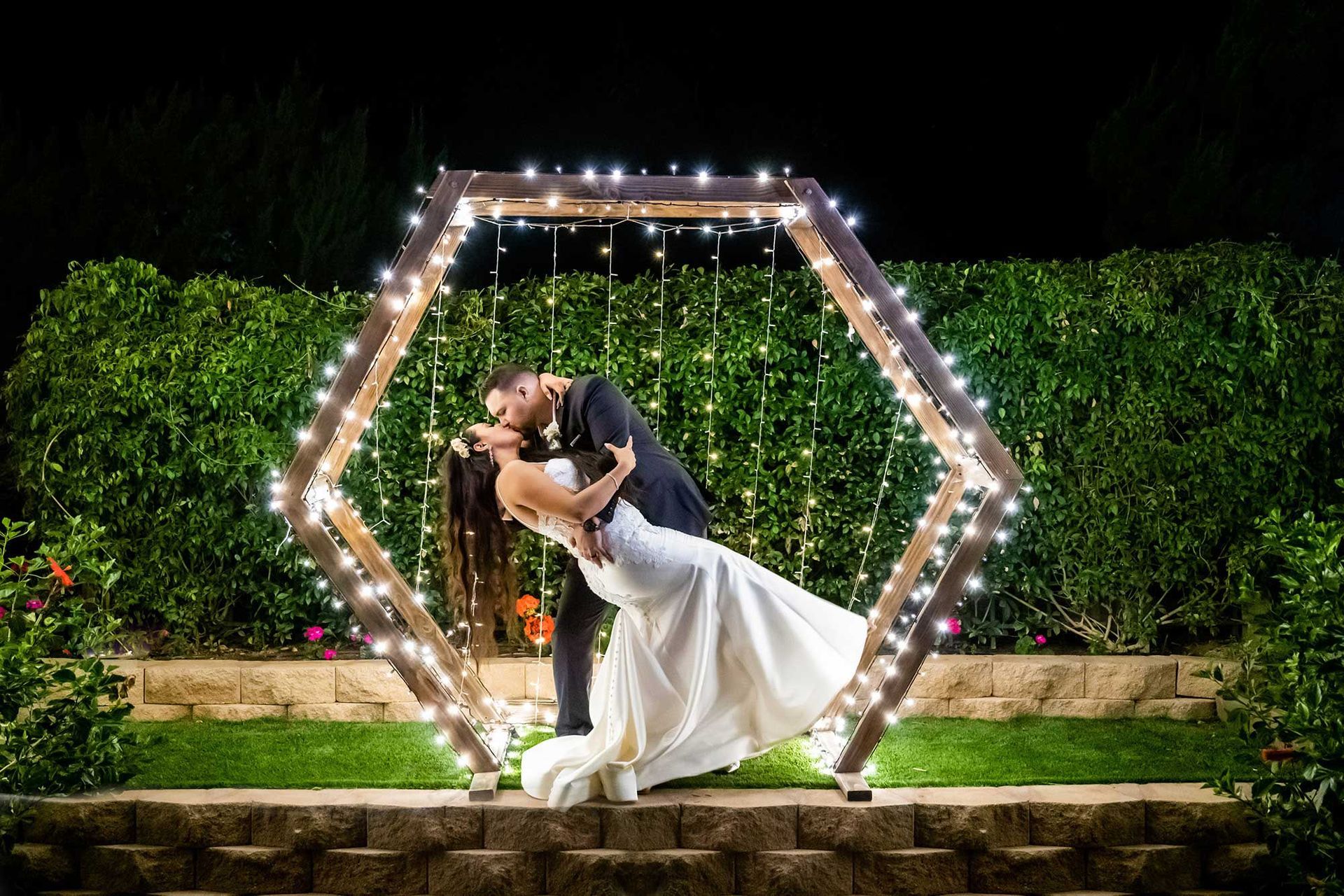 A bride and groom are kissing under a wooden arch with lights.