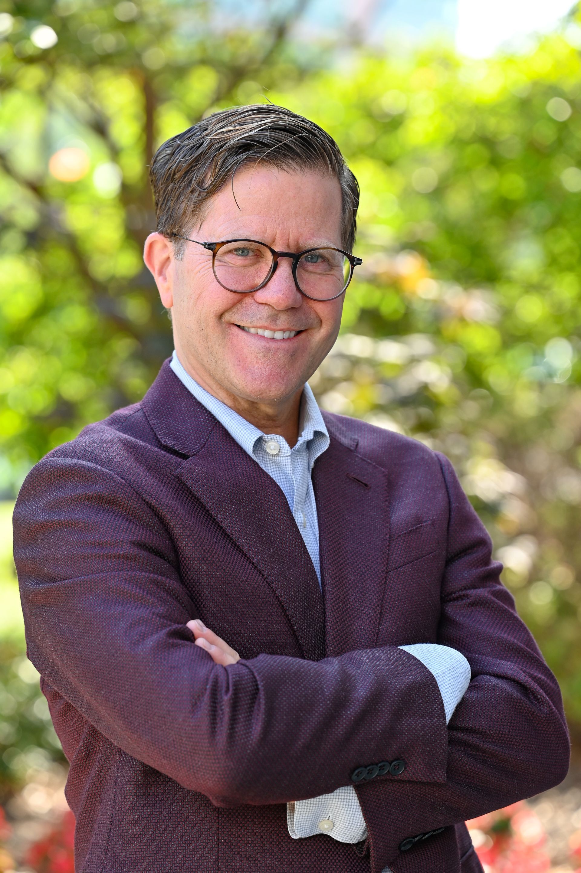 A man in a purple suit and glasses is smiling with his arms crossed.