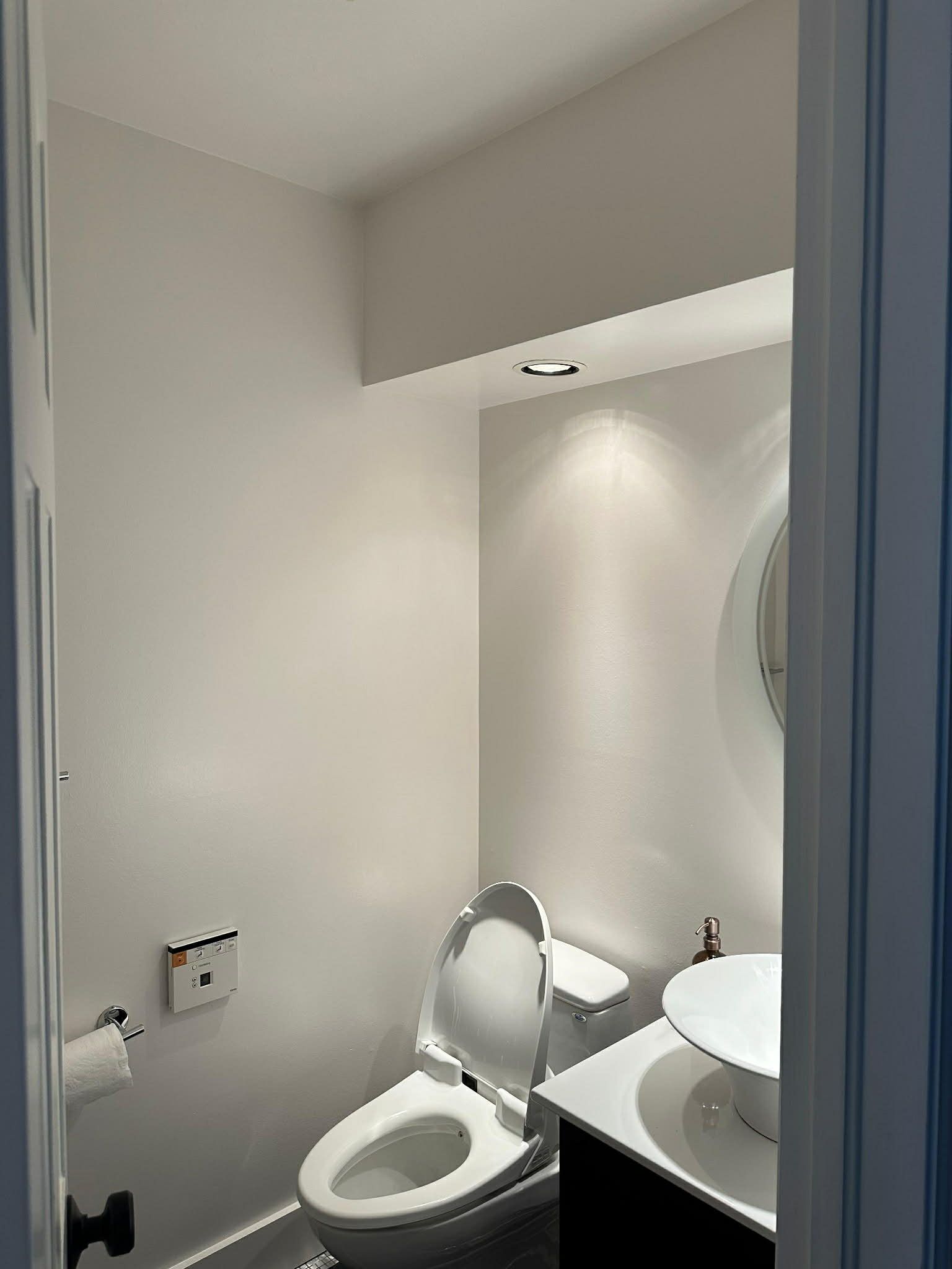 A modern, compact powder room with a white toilet, a dark vanity, a circular mirror, and recessed ceiling lighting.