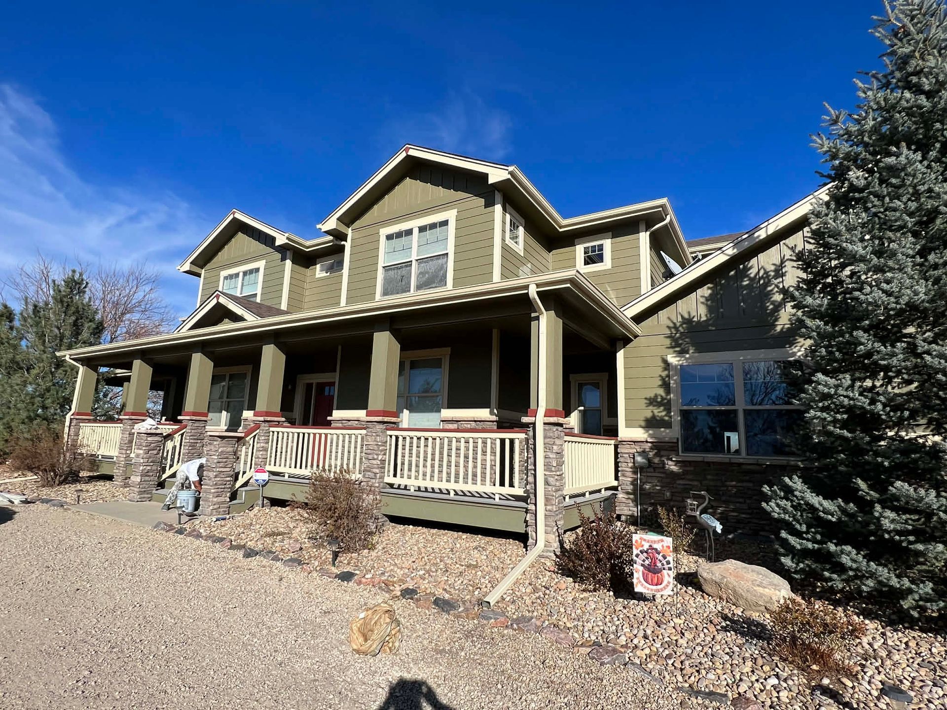 A two-story olive green house with a large front porch, stone accents, and a gravel yard under a clear blue sky.