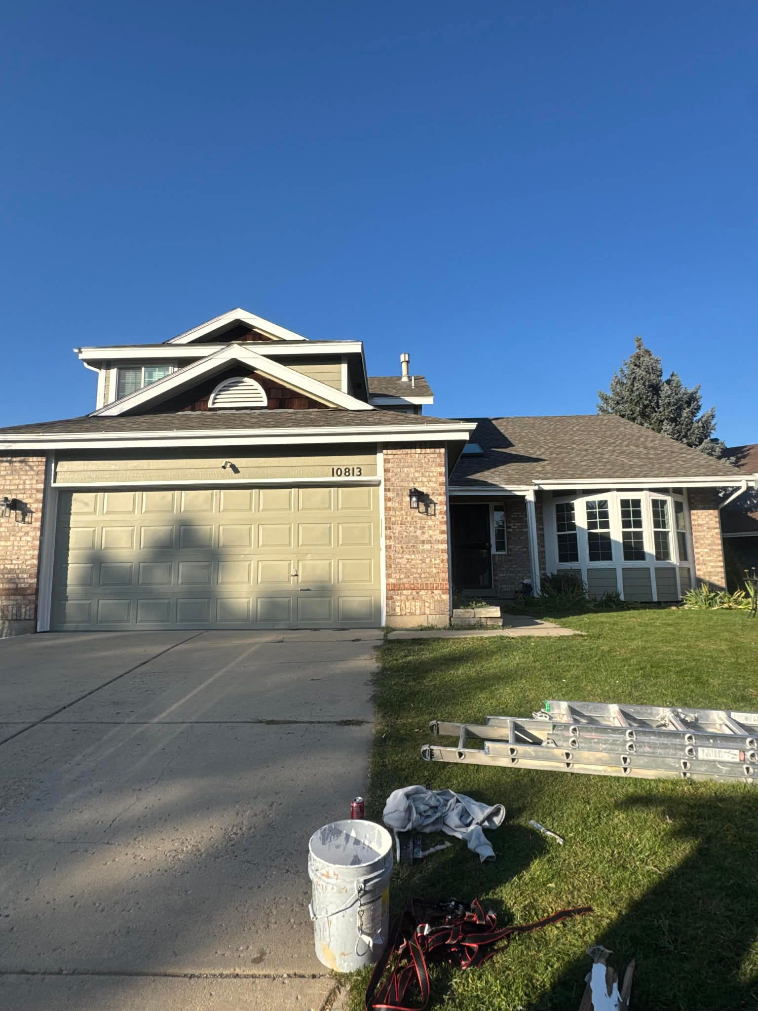 A two-story tan brick and siding house with a multi-gabled roof, garage, driveway, and lawn under a clear blue sky.